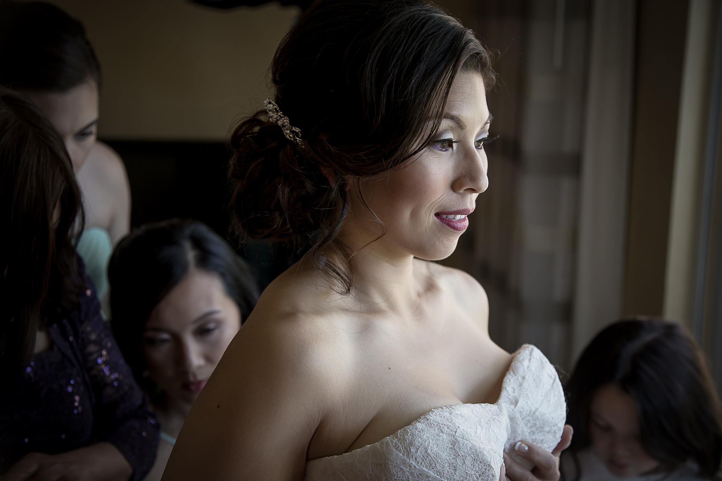 Bride with dark hair styled with a jeweled hair accessory wearing a strapless white lace wedding gown, surrounded by bridesmaids in dark dresses, as she gazes out a window.
