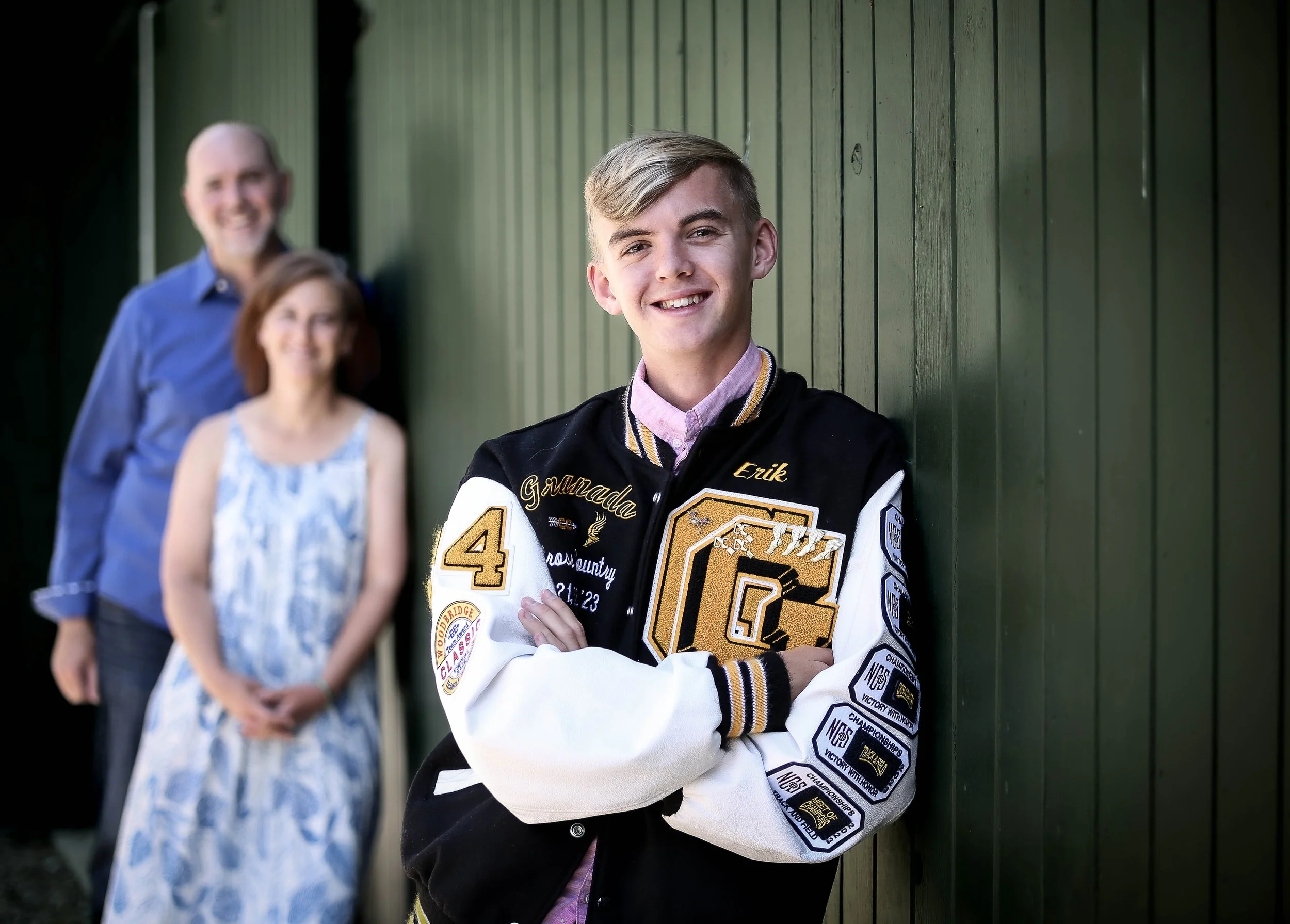 Three people standing in front of a green wooden wall. The young man in the foreground is smiling, wearing a black varsity jacket with patches and the name 'Erik'. Behind him, a man with glasses and a woman in a blue dress are slightly blurred.