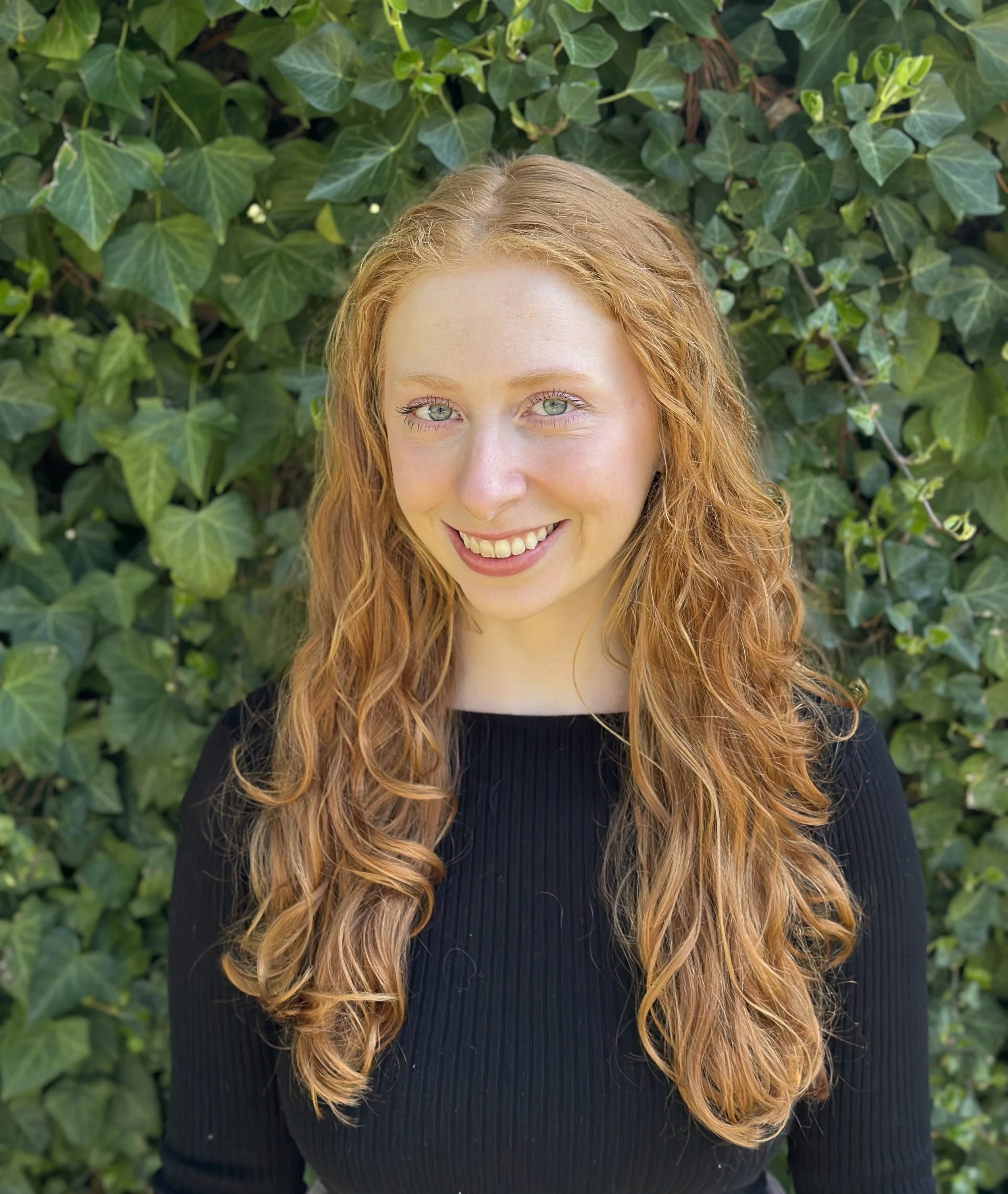 A young woman with curly red hair and blue eyes smiling in front of a leafy green background.