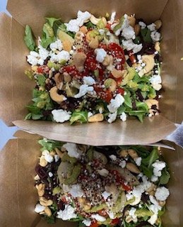 Two salads in brown takeout containers with lettuce, crumbled cheese, diced vegetables, and toppings.