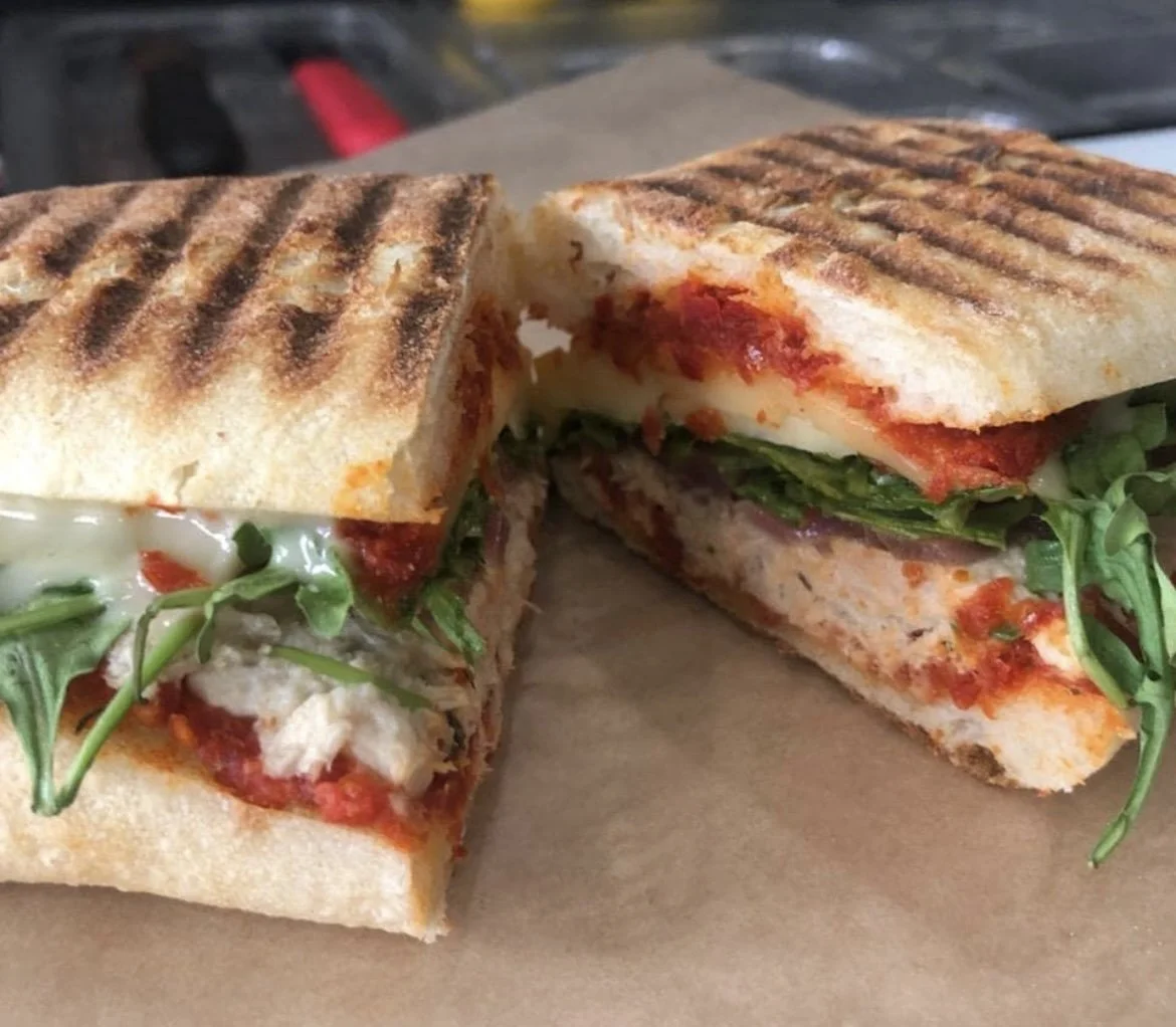 Close-up of a grilled sandwich cut in half, showing visible layers of melted cheese, tomato sauce, spinach, and chicken, with grill marks on the bread.