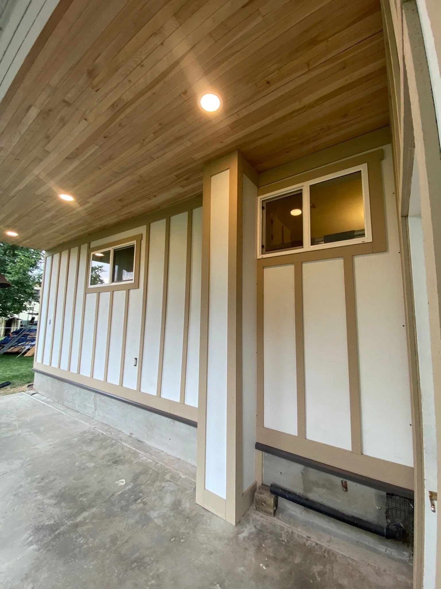 The image shows a partially finished exterior wall of a house with two small windows and a wooden ceiling with recessed lighting. The wall has vertical siding, and the floor appears to be concrete.