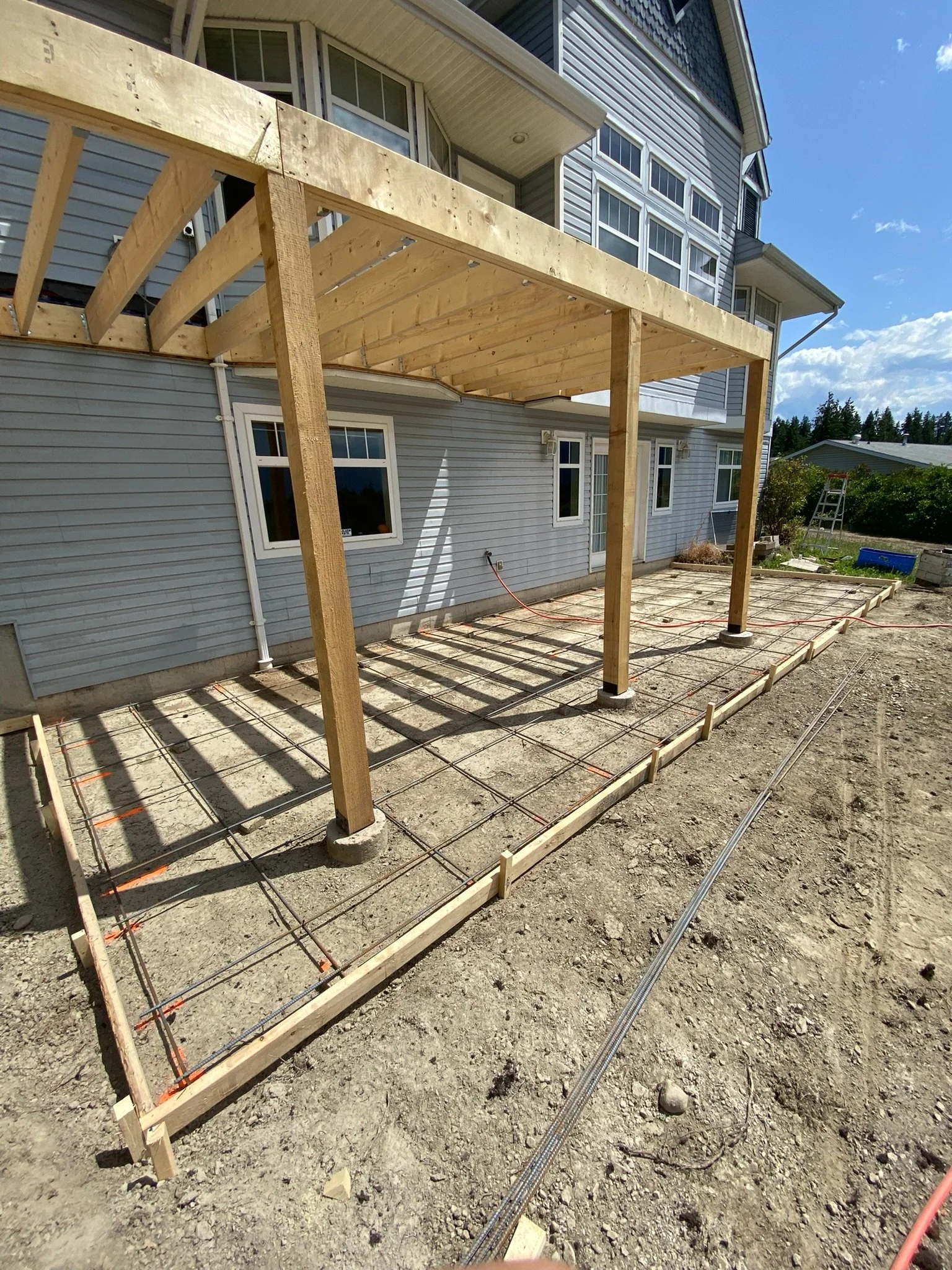 Construction site showing the framing of a deck attached to a blue house with gray siding and multiple windows, with a ladder and tools in the background under a partly cloudy sky.