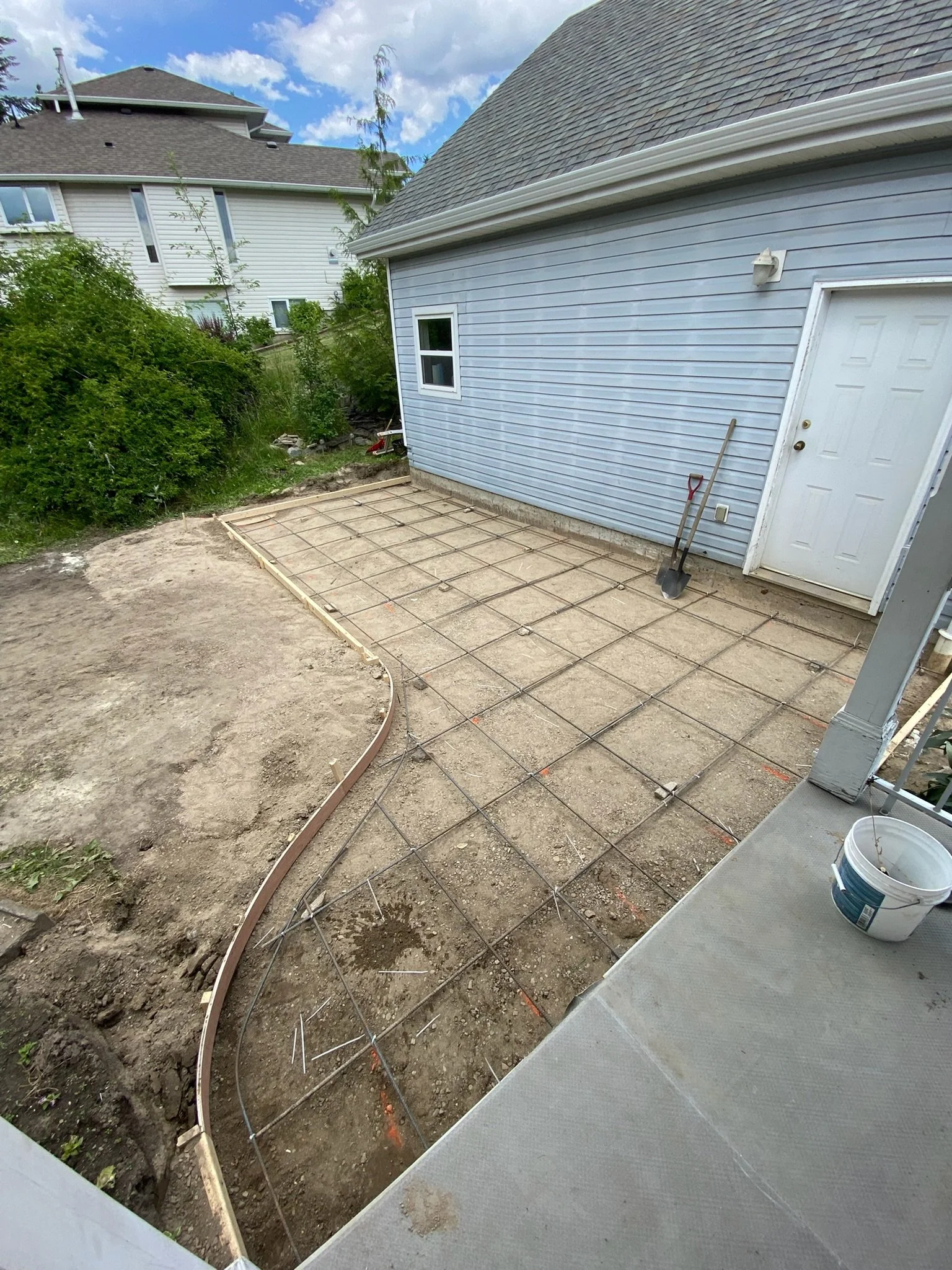 A backyard where a concrete patio is being constructed with reinforced steel grid in place. The surrounding area shows dirt and construction tools, including a shovel and piping, near a light blue house with white trim and a gray shingled roof.