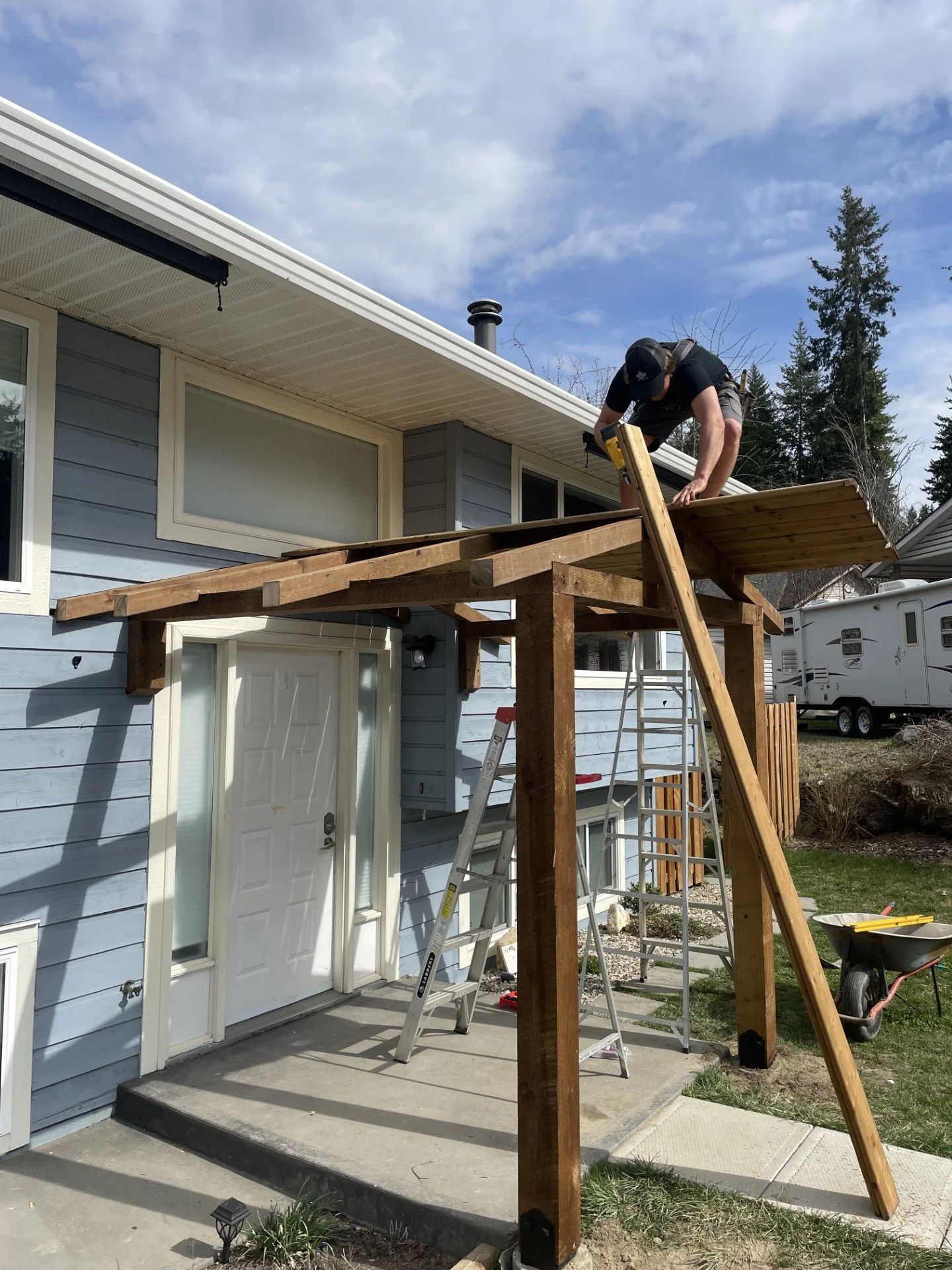 A person standing on a ladder and working on a wooden deck extension outside a light blue house with a white door and windows, under a partly cloudy sky.
