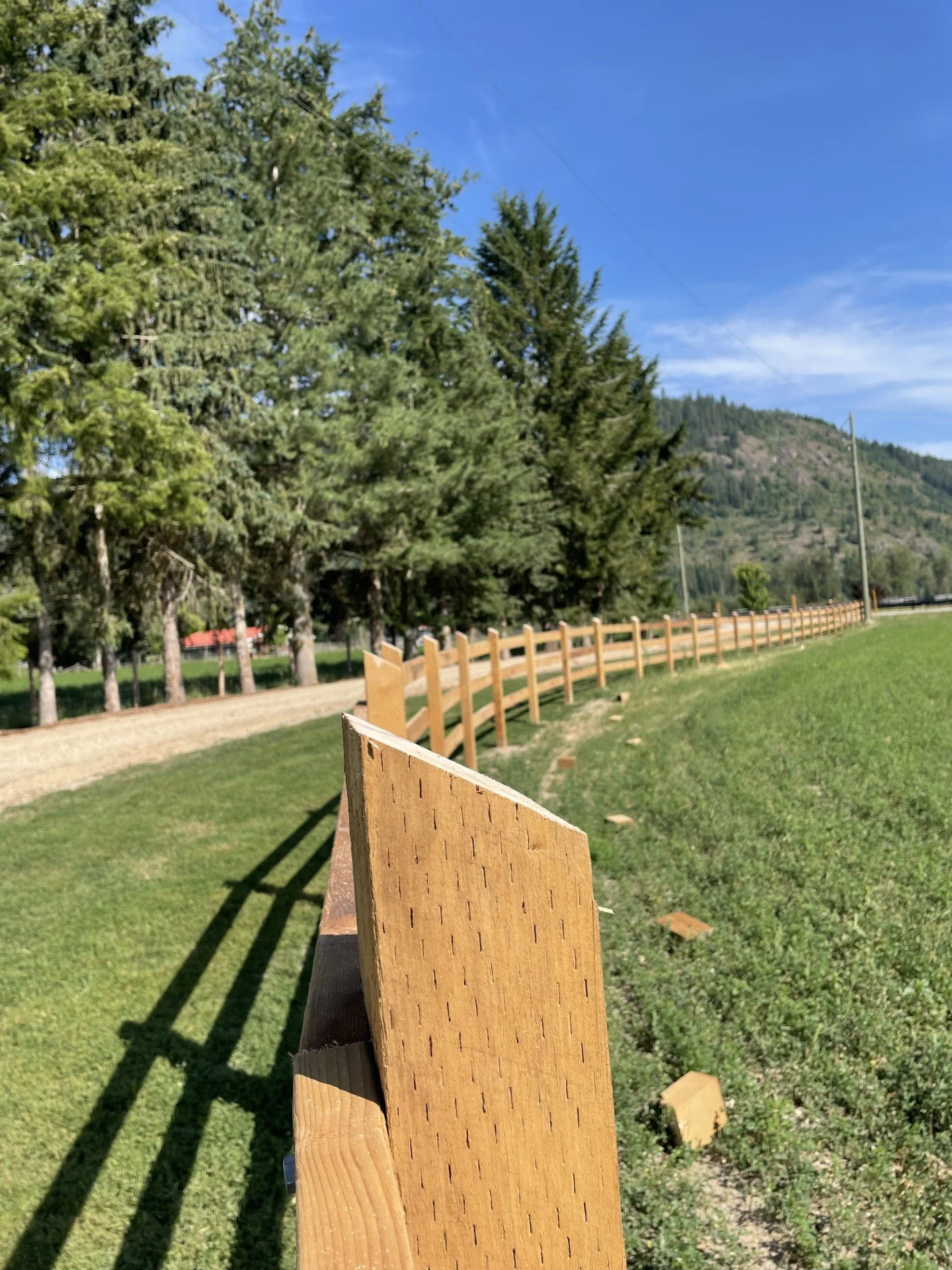 A wooden fence running along a grassy lawn with trees and mountains in the background, under a blue sky with some clouds.