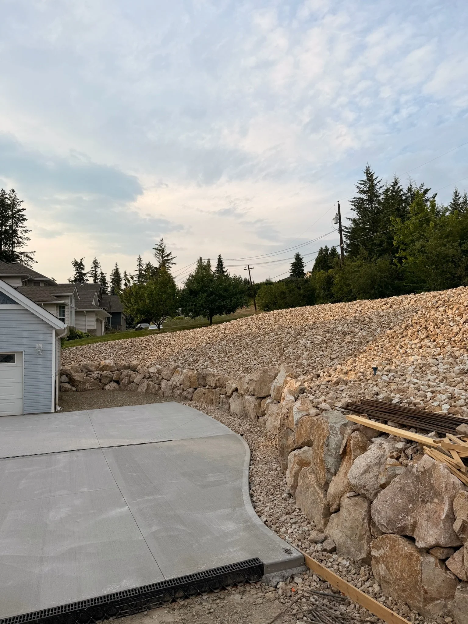 Concrete driveway next to a rock wall with house and trees in the background.