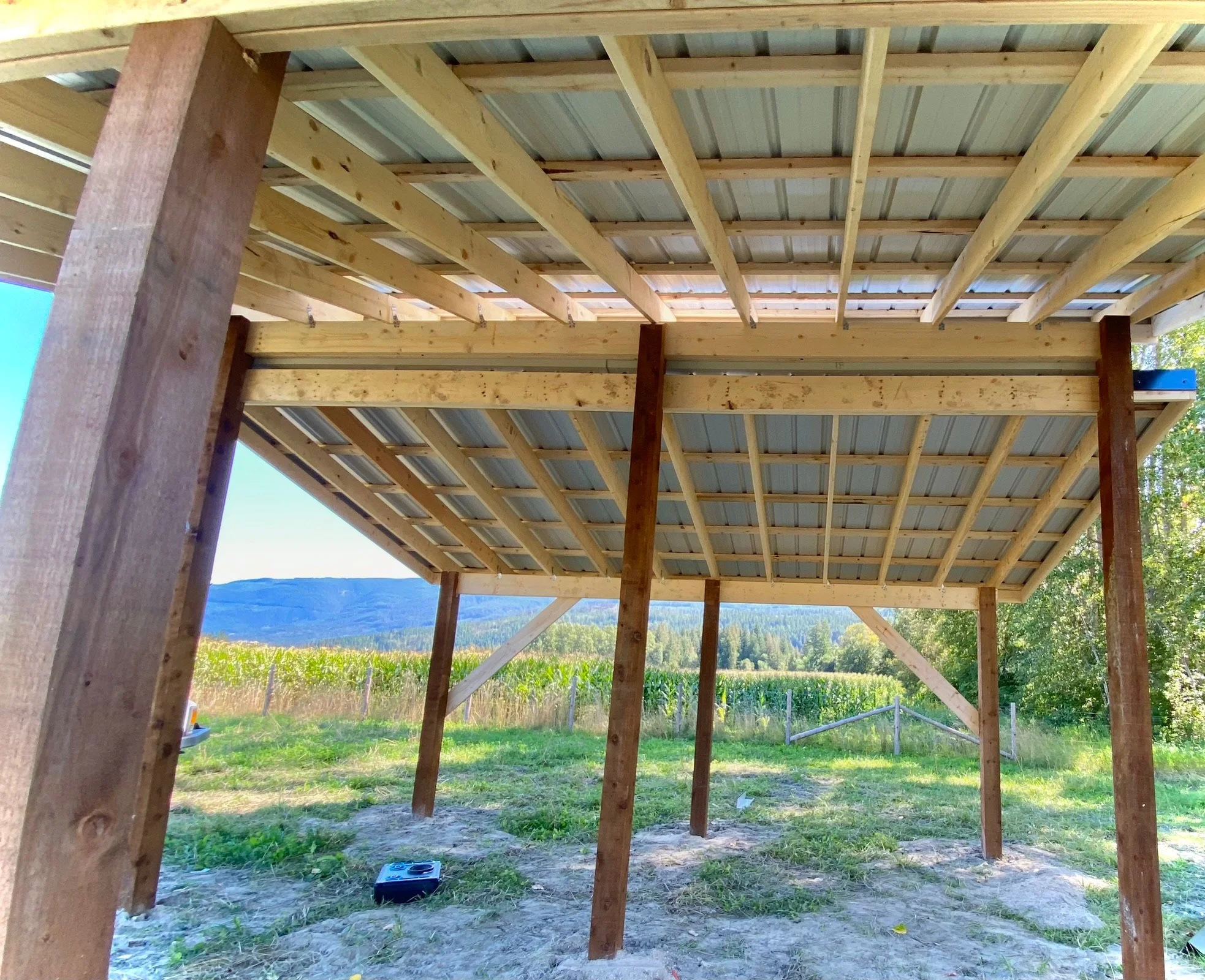 Construction site showing a wooden structure with a corrugated metal roof frame in a rural area with green fields and mountains in the background.