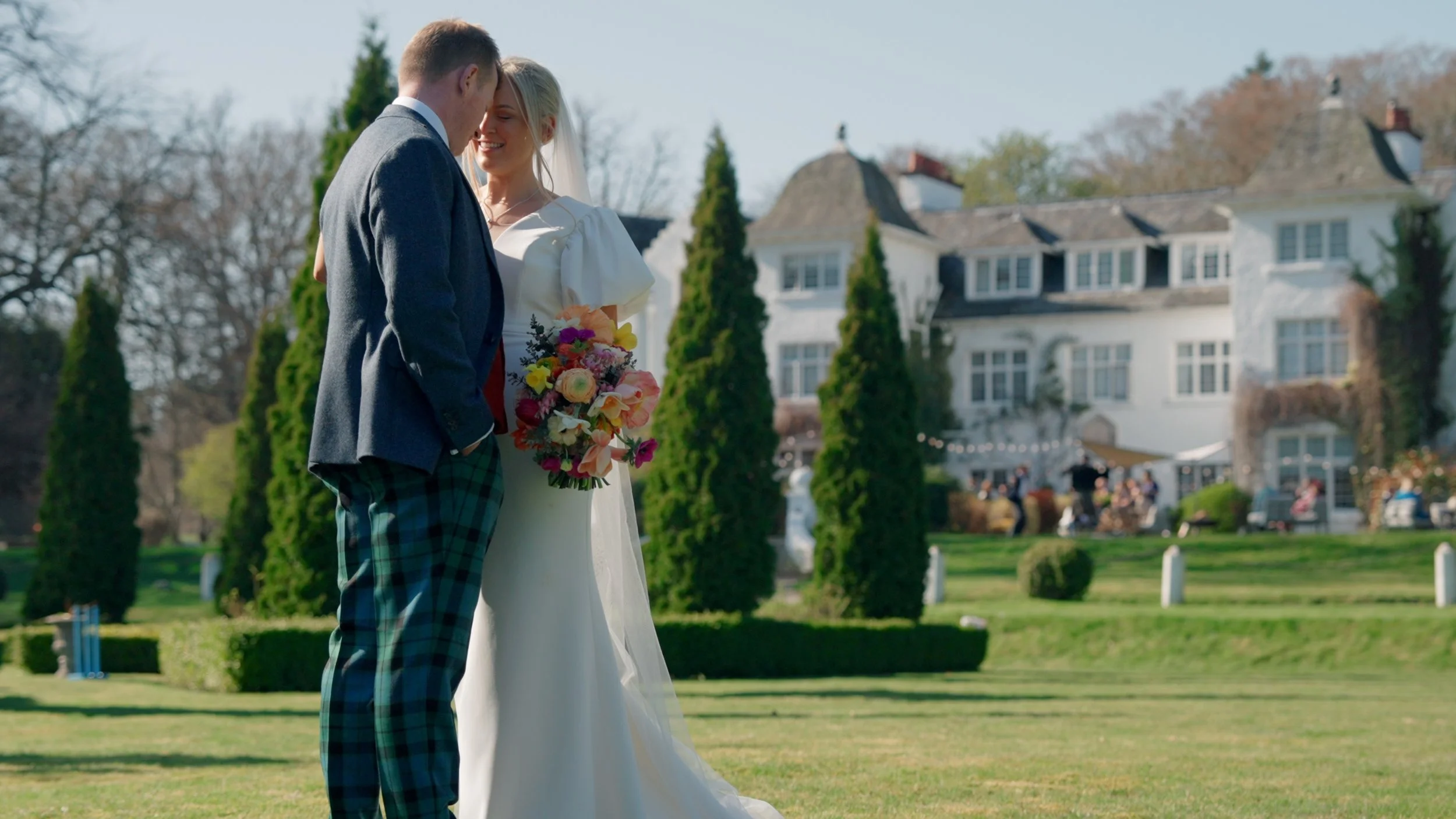 A bride and groom stand close together holding hands and smiling, outdoors on a bright day with a large white house and trees in the background.