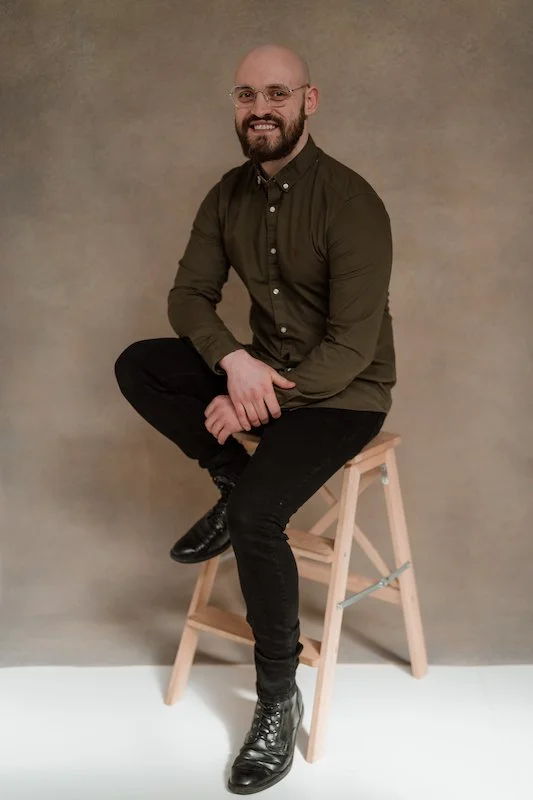 A man with a bald head and beard, wearing glasses, a dark button-up shirt, black pants, and black boots, sitting on a wooden stool against a plain background, smiling at the camera.