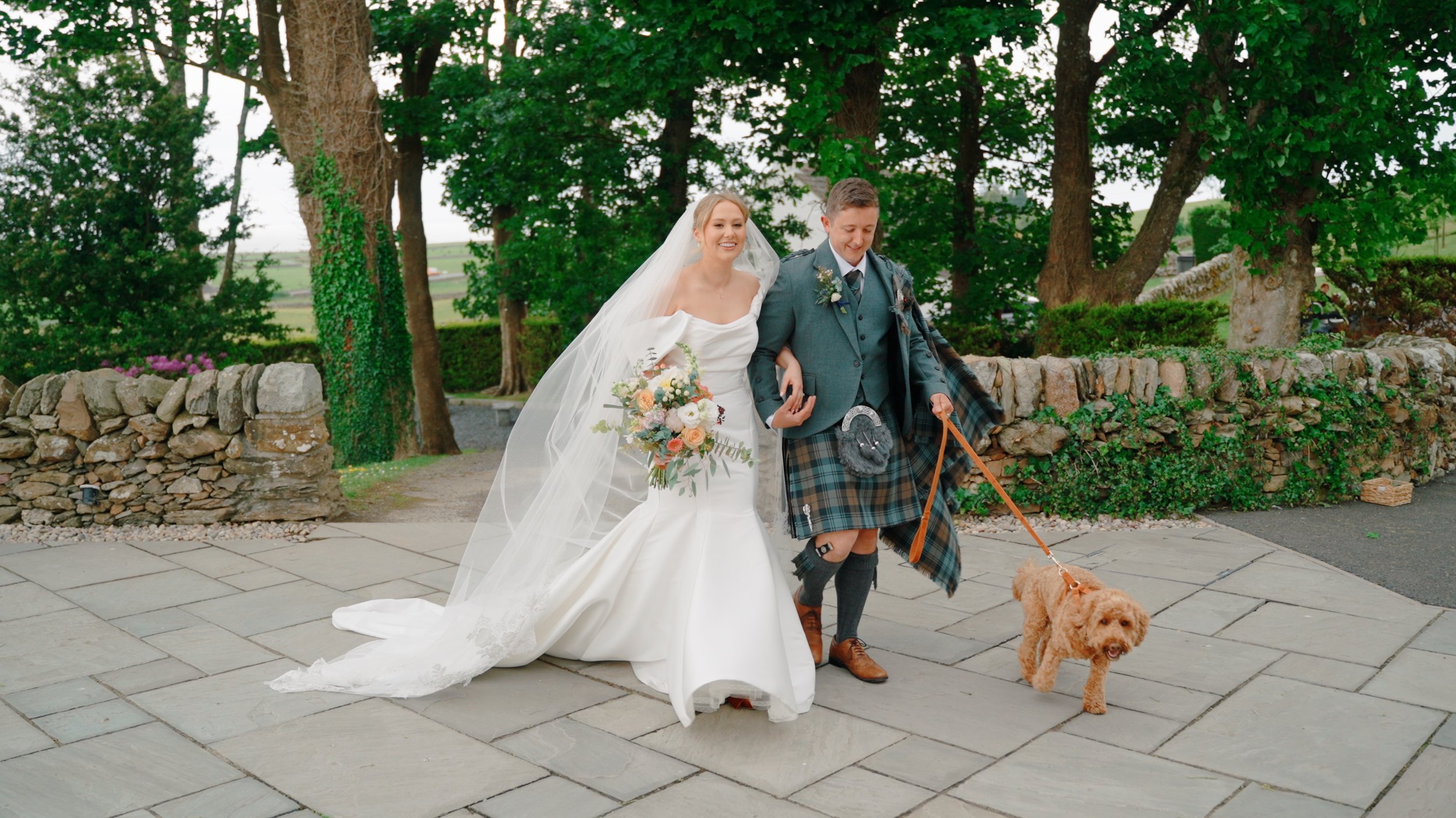 A bride in a white wedding dress walking arm-in-arm with a man in traditional Scottish attire, including a kilt, while walking two dogs outdoors on a paved area with trees and a stone fence in the background.