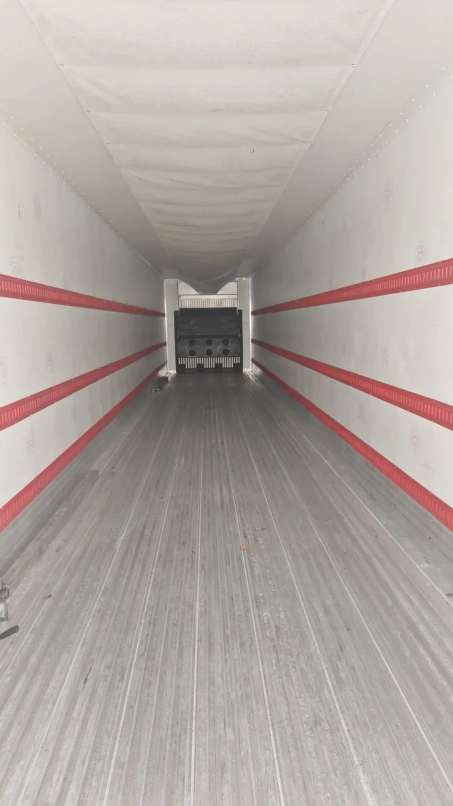 Interior de un remolque de camión con paredes blancas, líneas rojas y suelo de madera, con la puerta trasera abierta.