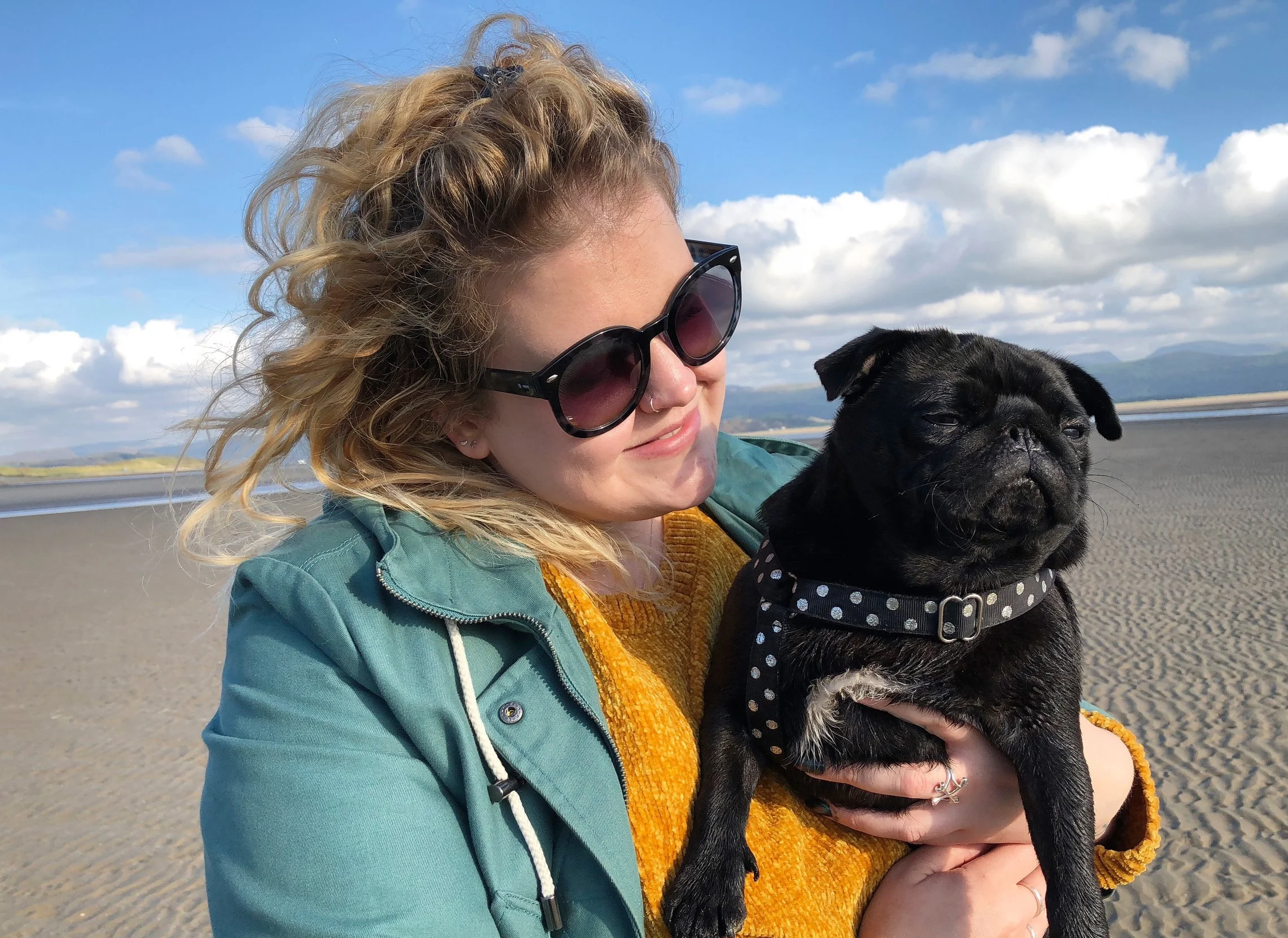 A woman with curly blonde hair wearing large black sunglasses, a green hoodie, and a yellow sweater, holding a black French Bulldog puppy with a polka dot collar against a backdrop of a cloudy blue sky and open landscape.
