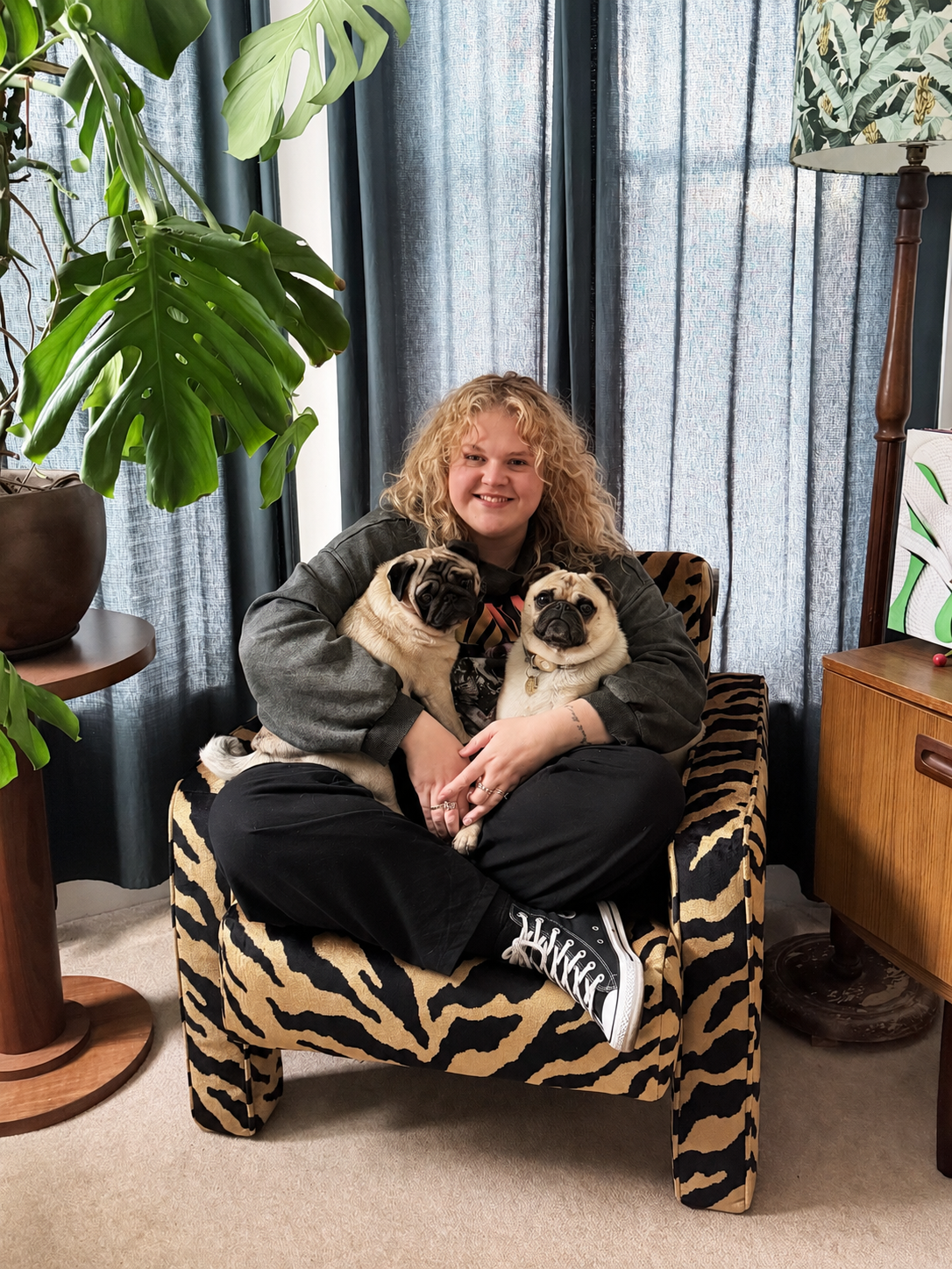 A woman with curly blonde hair sitting on a Tiger print armchair, holding two pug dogs, in a living room with plants and blue curtains.