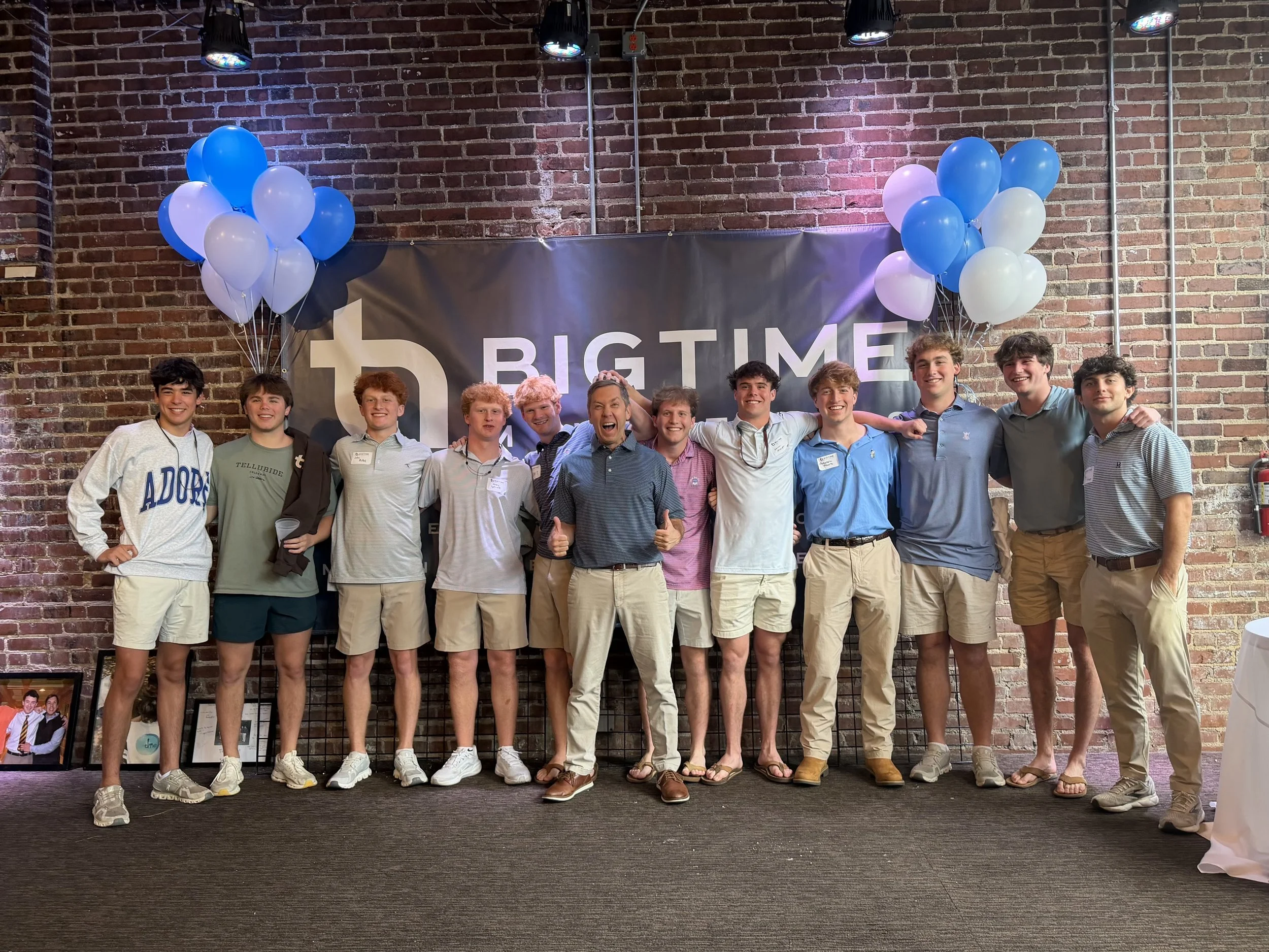 Group of young men and women standing together in front of a brick wall at an event with blue and white balloons on each side and a black banner that reads 'Big Time' in the background.