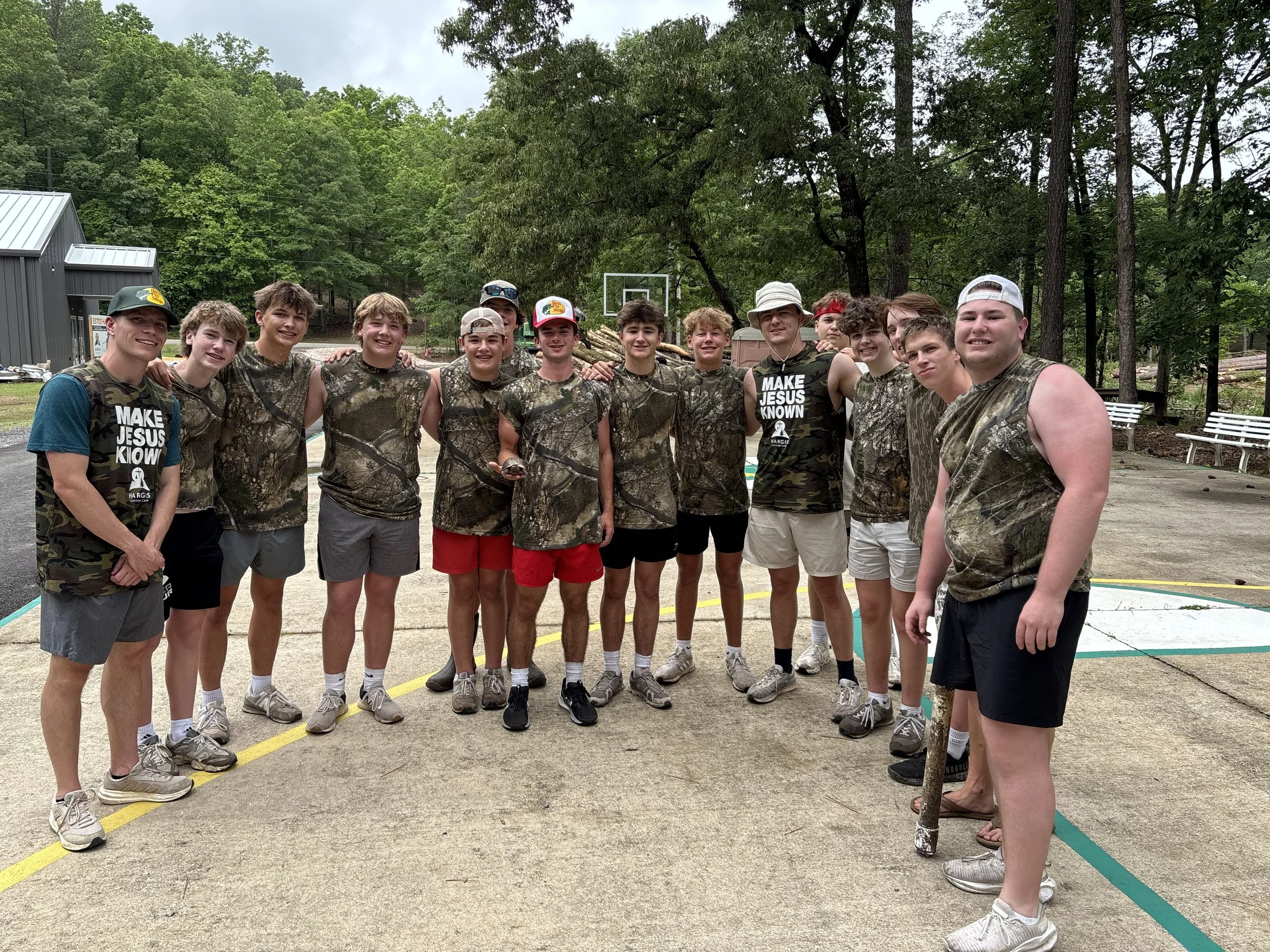 A group of boys and young men standing outdoors on a concrete court, all wearing camouflage clothing, with some smiling and posing for the photo. They are surrounded by trees and outdoor sports equipment.