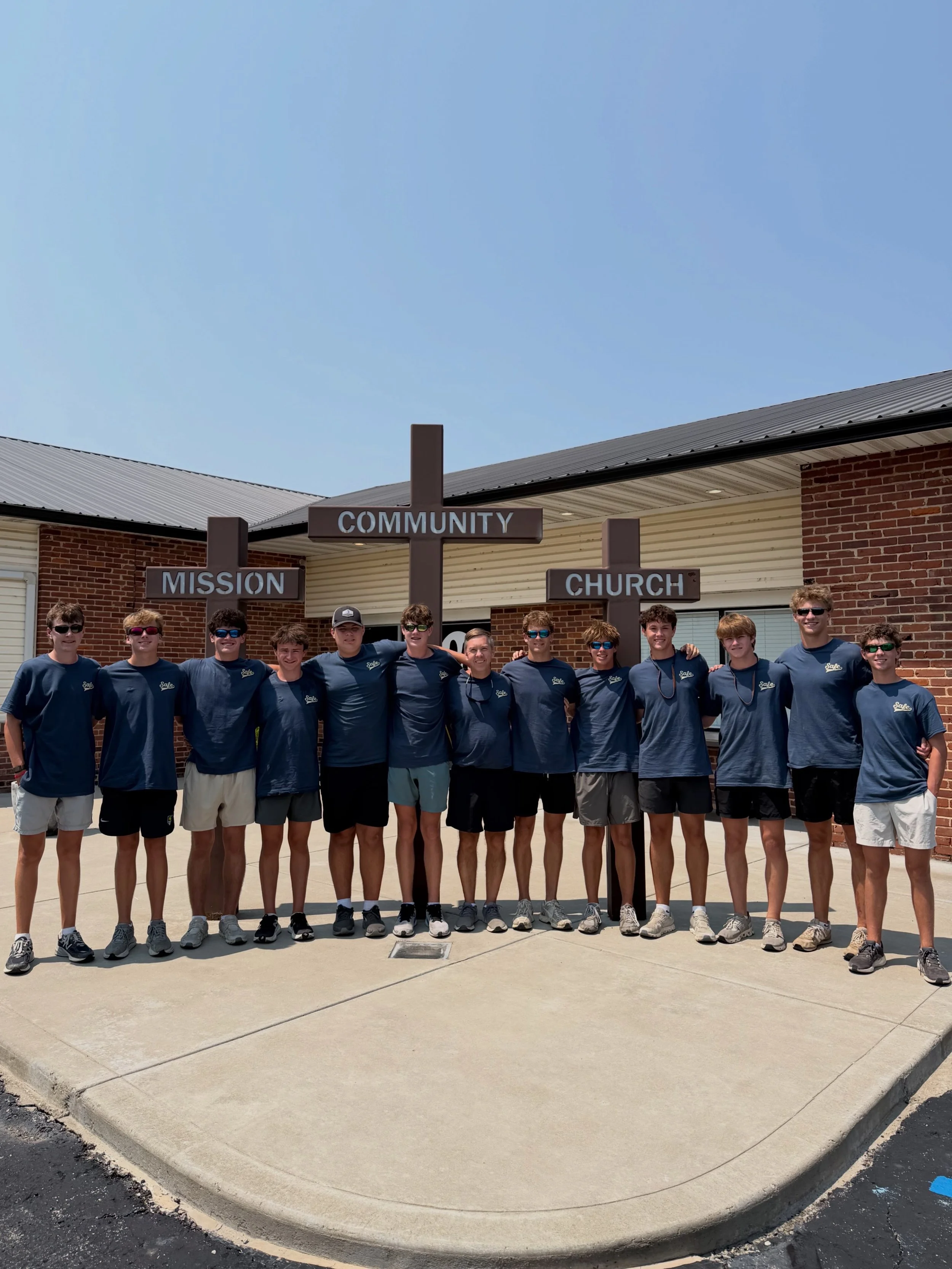 Group of young men standing outside in front of a brick building with signage that reads 'Mission', 'Community', and 'Church'. They are smiling and posing for a group photo, all wearing matching blue T-shirts and sunglasses.