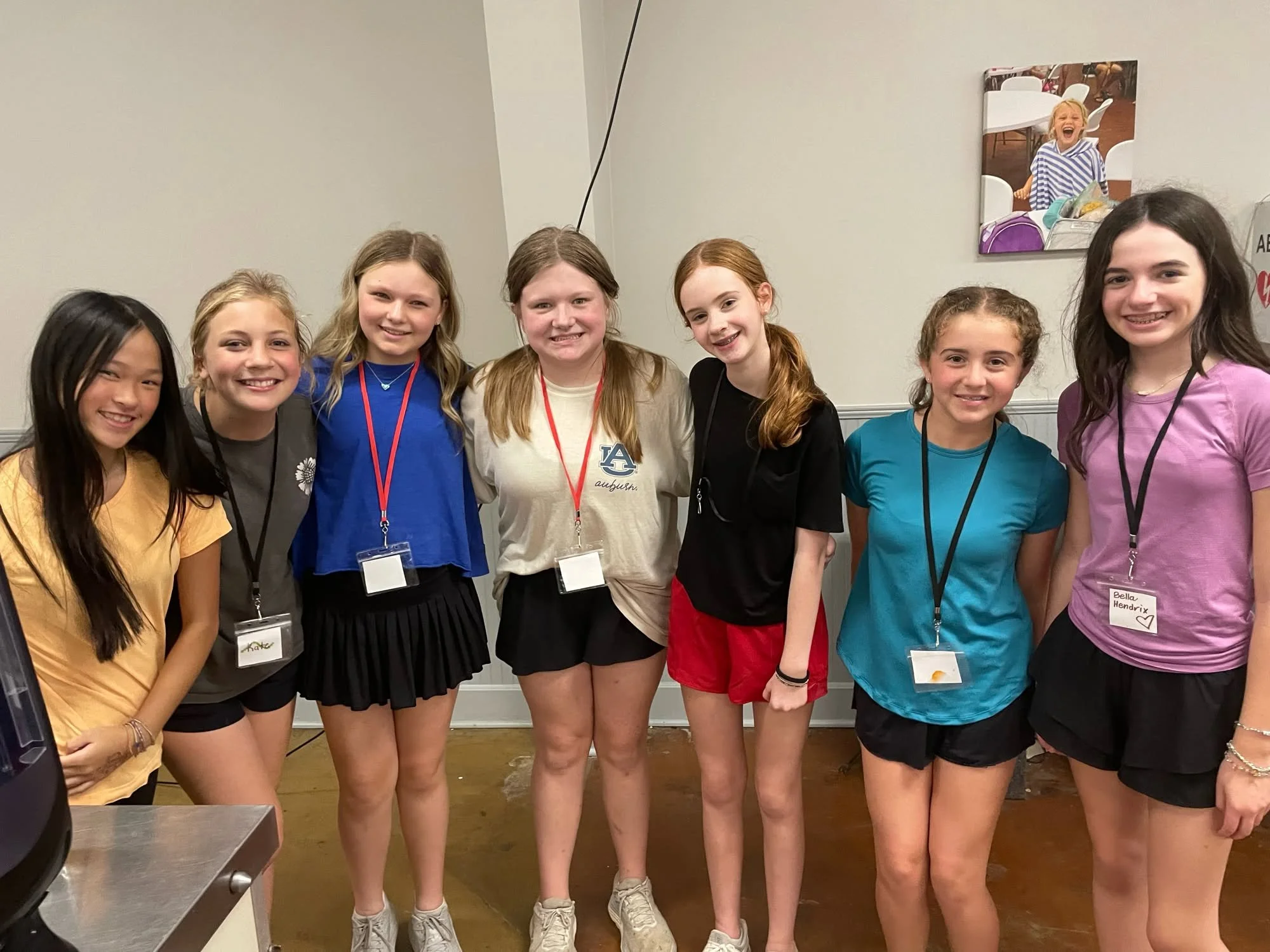 Group of eight young girls standing together indoors, smiling for the photo. They are wearing casual clothing and name tags, with some wearing shorts and t-shirts. One girl has a white Auburn University shirt.