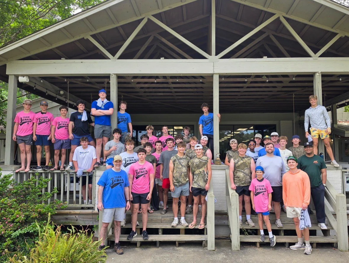 A large group of teenagers and young adults posing together on the steps and porch of a wooden building, some wearing summer clothes and team T-shirts that say 'Team Jesus'.