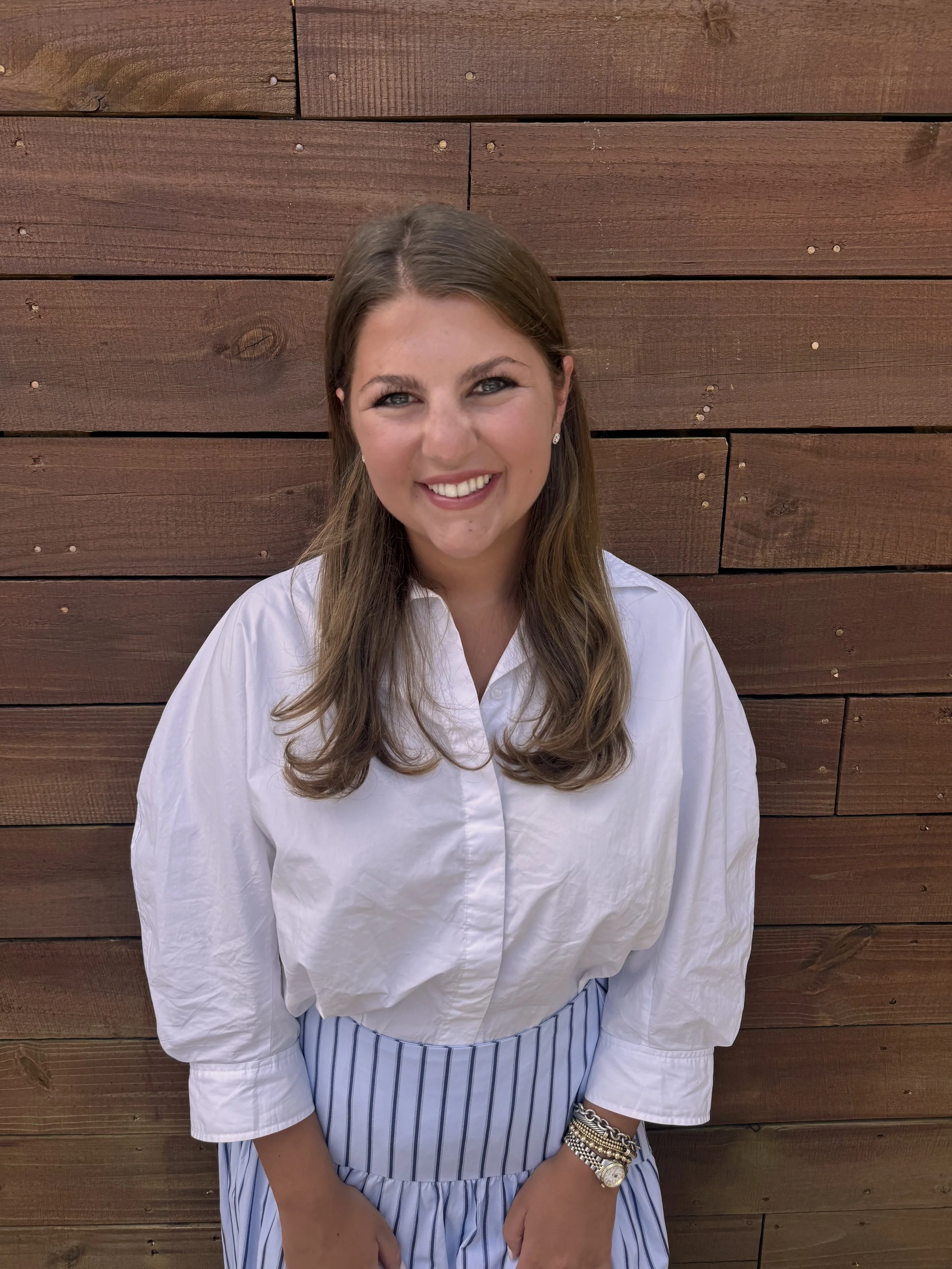 A young woman with long brown hair and blue eyes, smiling, wearing a white blouse, striped skirt, and jewelry, standing against a wooden wall.