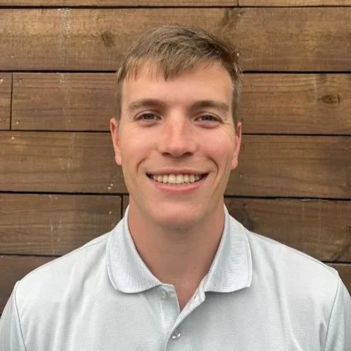 A young man with light brown hair and a big smile, wearing a white collared shirt, standing in front of a wooden background.