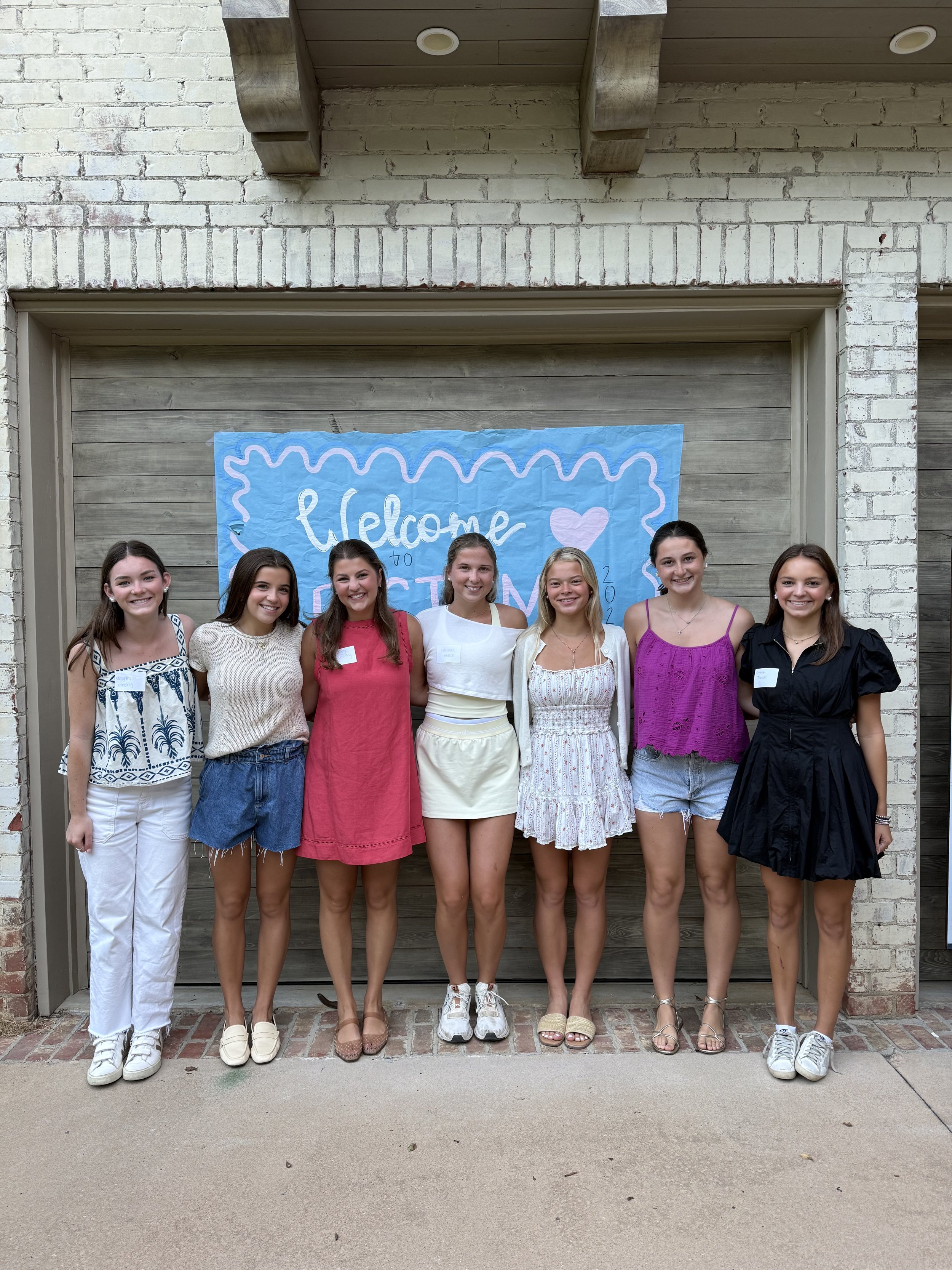 Group of seven young women standing in front of a brick and wood wall with a blue 'Welcome' banner, dressed in summer clothing, smiling at the camera.