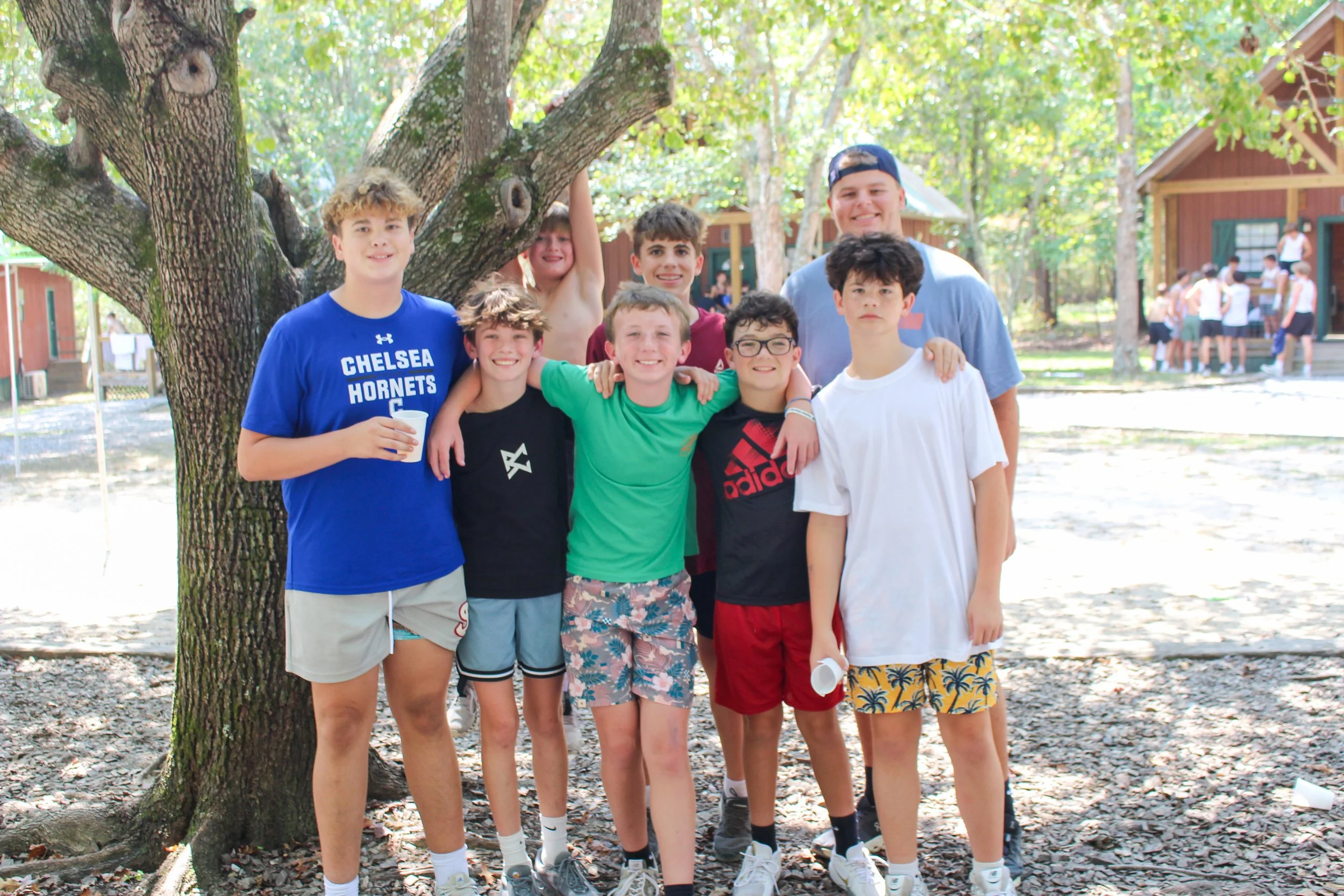 A group of ten boys standing together outdoors next to a tree, smiling at the camera. They are in a wooded area, with wooden cabins in the background, likely at a summer camp.
