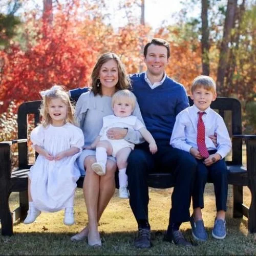 Family of five sitting on a park bench with fall foliage behind them. The parents are smiling, with two daughters and a son, all dressed in nice clothes.