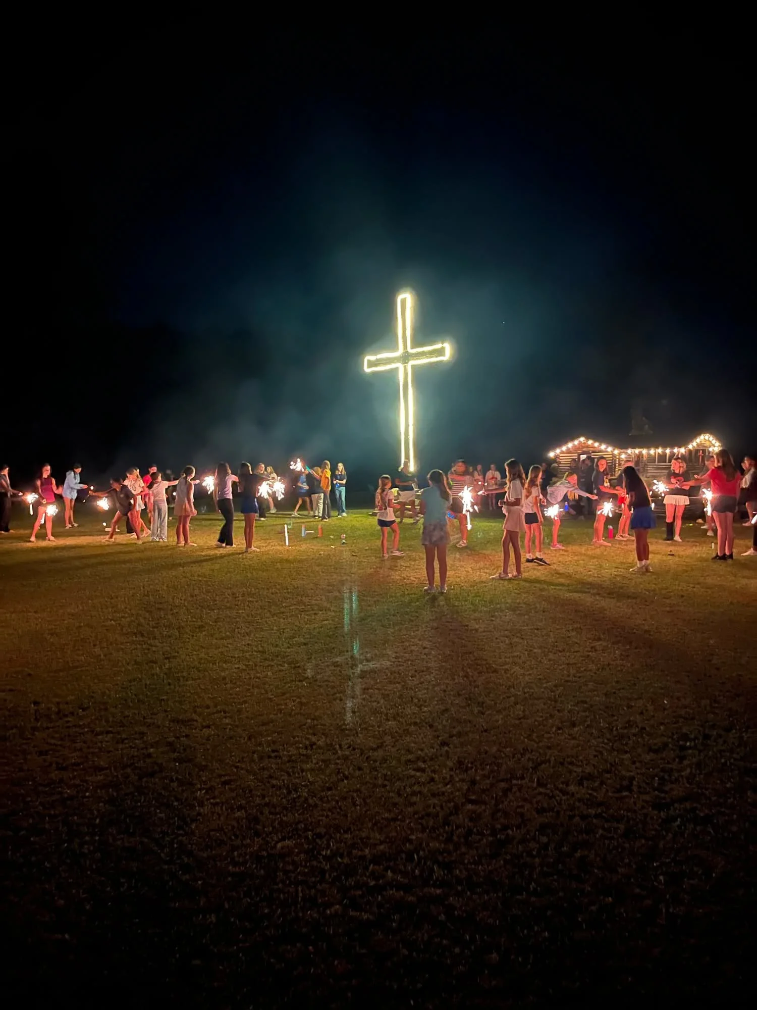 Nighttime outdoor scene with a large illuminated cross and a group of people holding sparklers, celebrating around a lit wooden structure in the background.