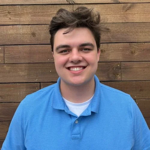 A young man with brown hair, smiling, wearing a blue polo shirt, standing in front of a wooden fence.