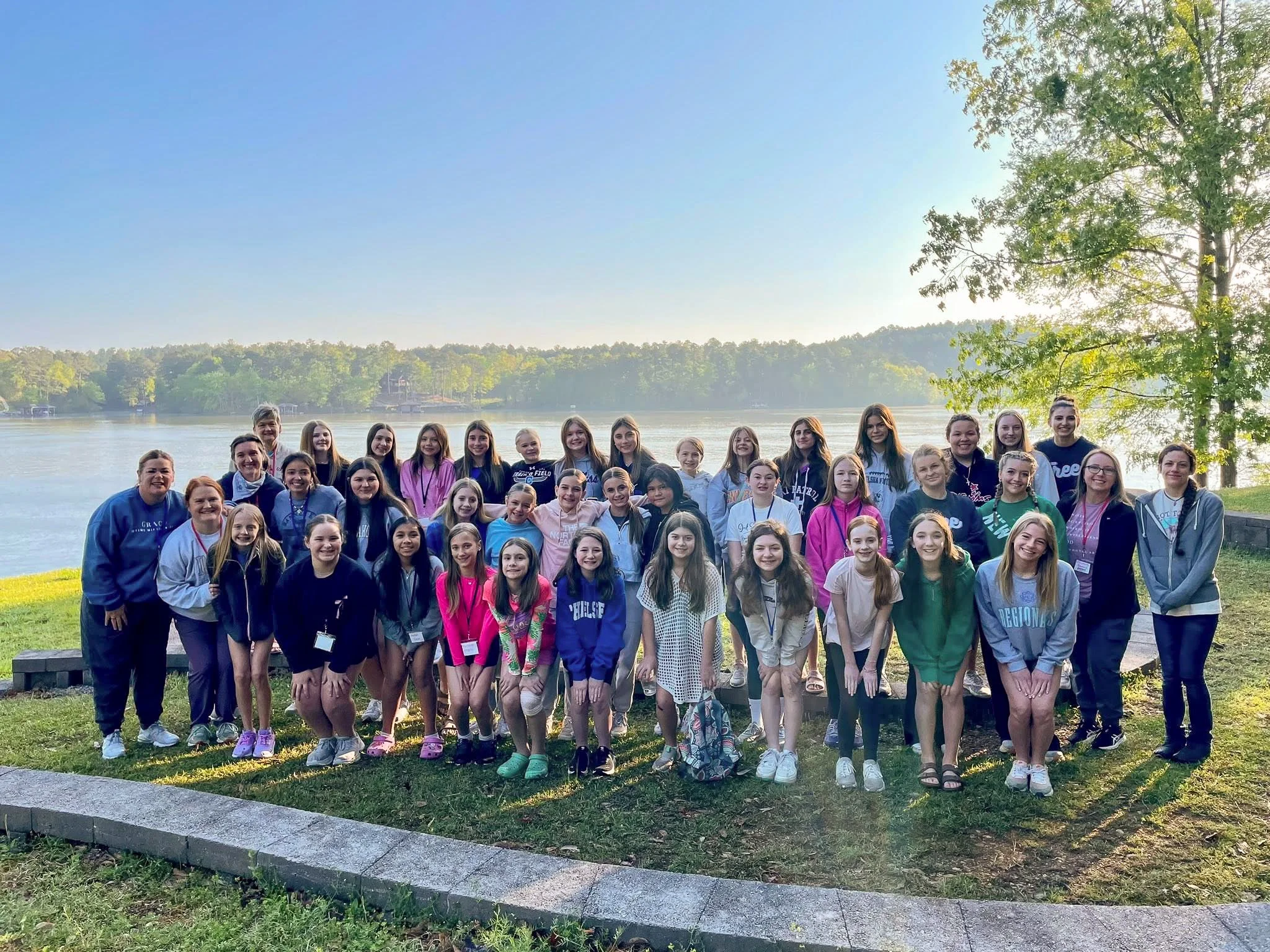 A large group of young girls and women posing together outdoors near a body of water on a sunny day.