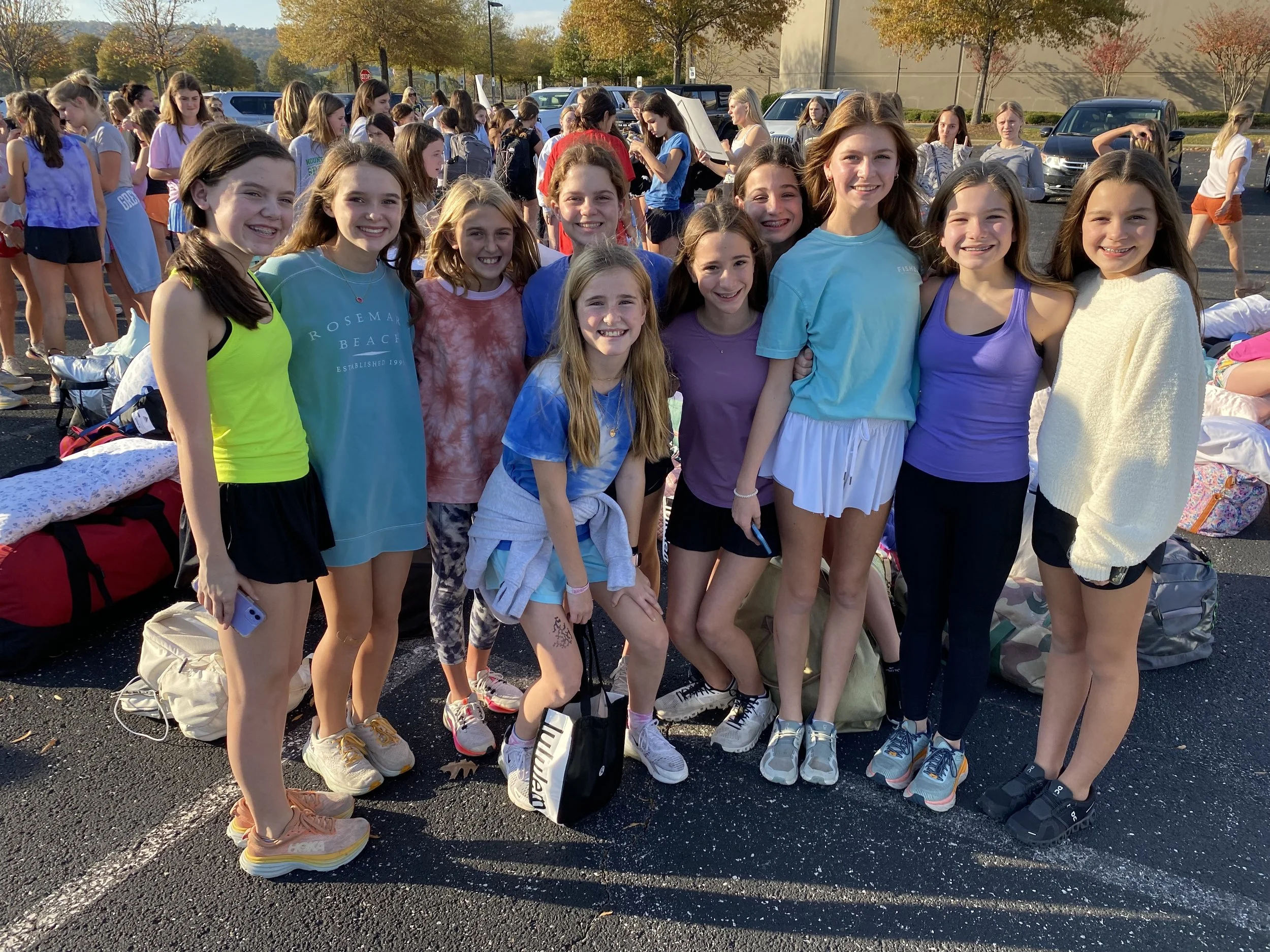 Group of schoolgirls smiling and standing together outdoors, with backpacks on the ground, during daytime with autumn trees in the background.