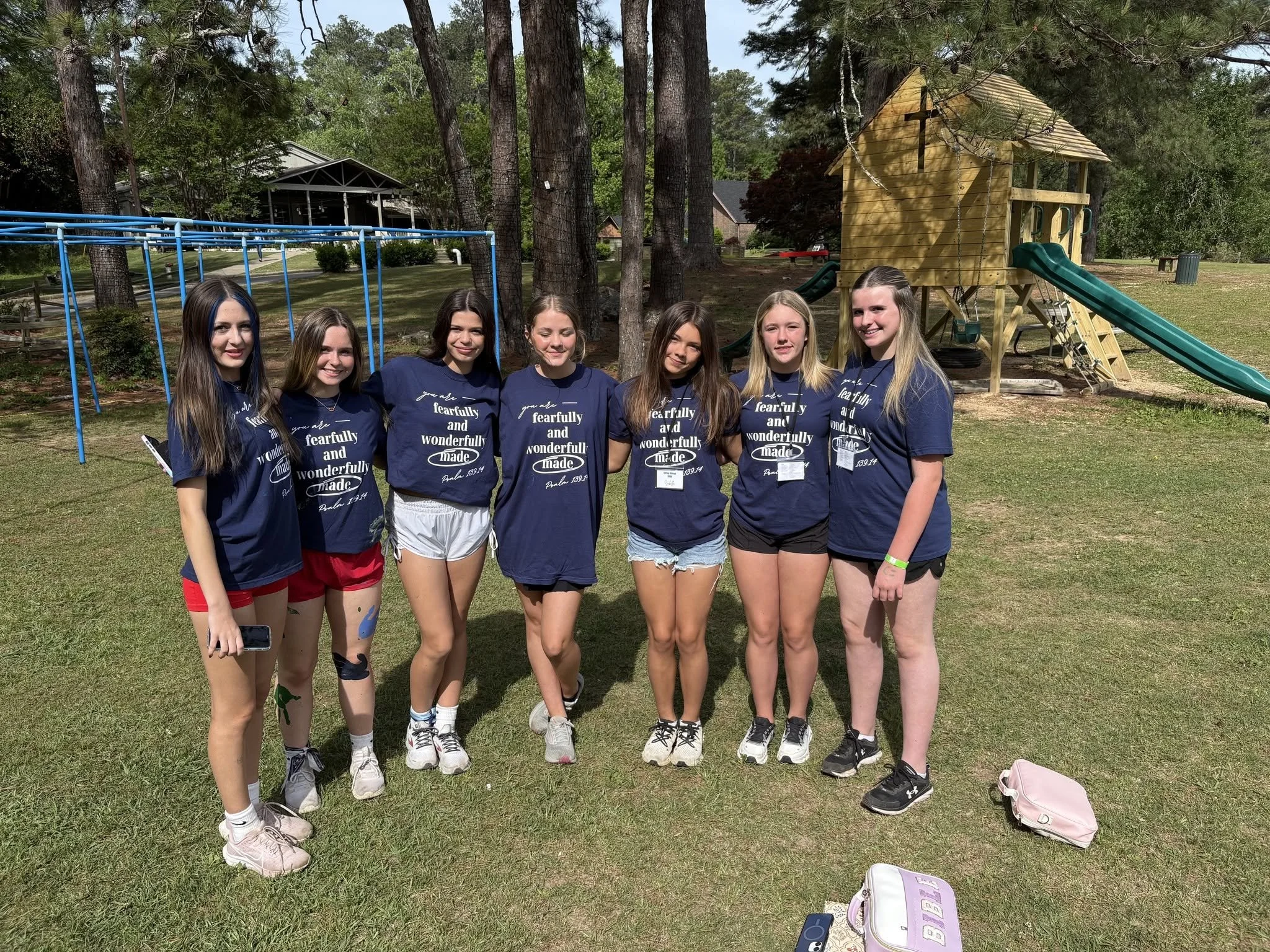 Seven girls standing outdoors in front of a wooden playhouse and a blue swing set, smiling at the camera. They are wearing matching navy blue t-shirts with white text, and shorts or skirts. The setting is a grassy area with tall trees and a few houses in the background.