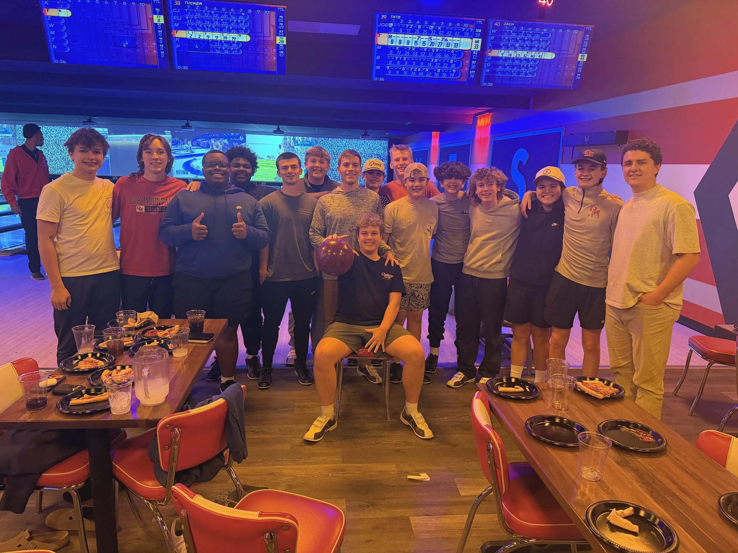 Group of boys and teenagers at a bowling alley, some smiling and posing for the photo, with bowling scores on screens above and pizza, drinks on tables.