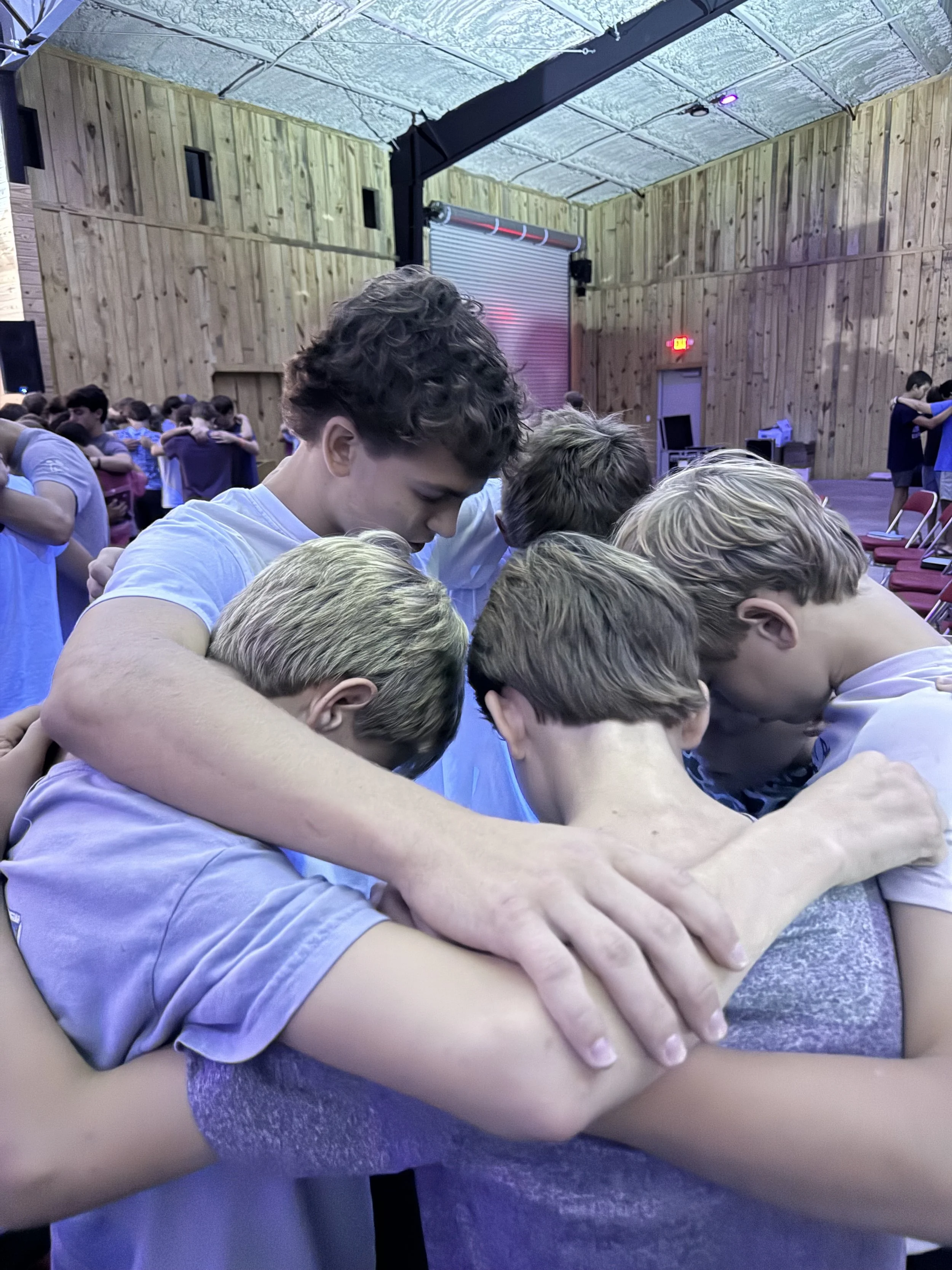 A group of people, mostly young males, are standing close together with their heads bowed and arms around each other in a circle inside a wooden-paneled room, possibly praying or participating in a group activity.