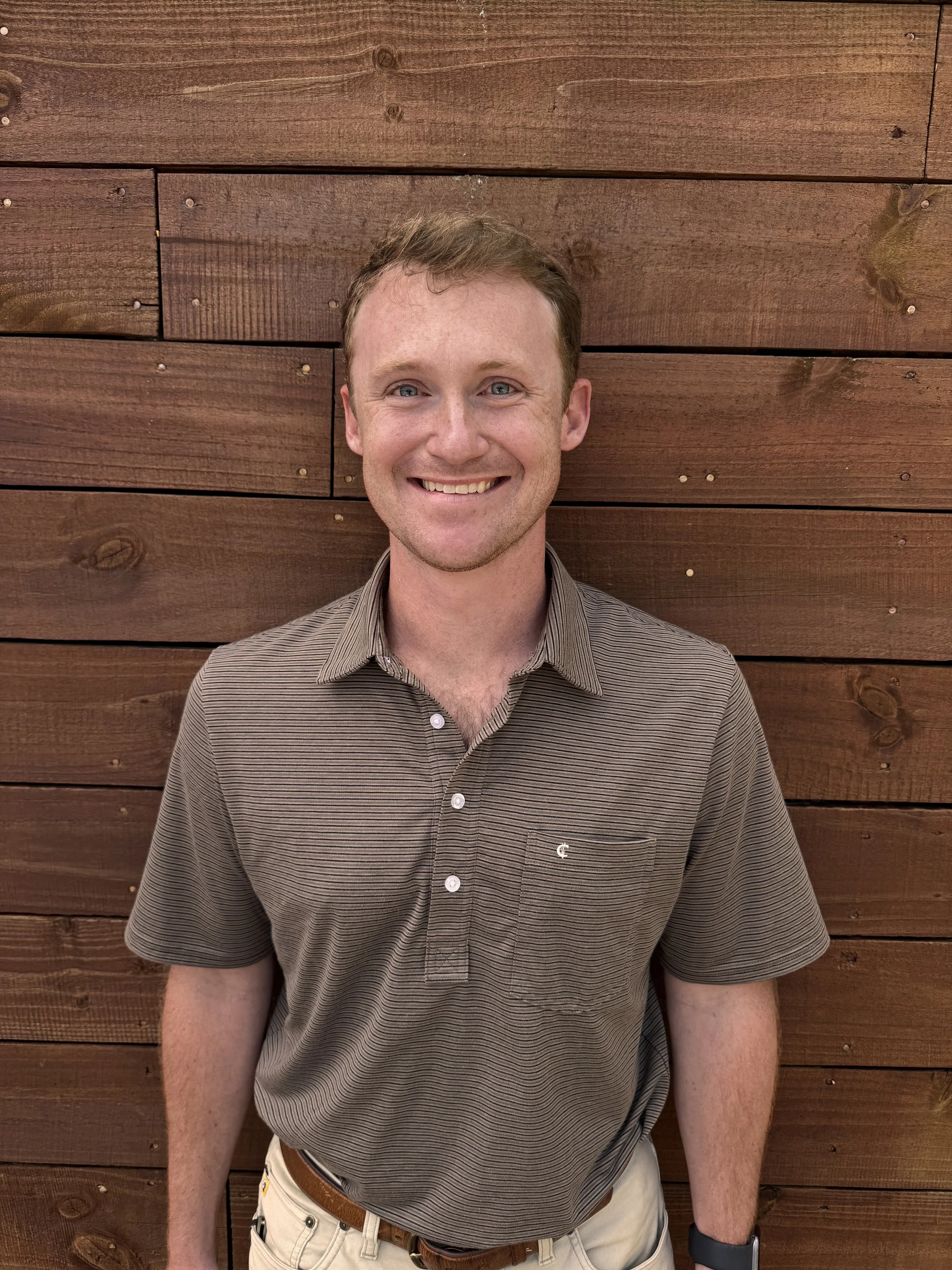 A young man with short light brown hair, blue eyes, and light skin, smiling, wearing a brown striped polo shirt and beige pants, standing in front of a wooden wall with horizontal planks.
