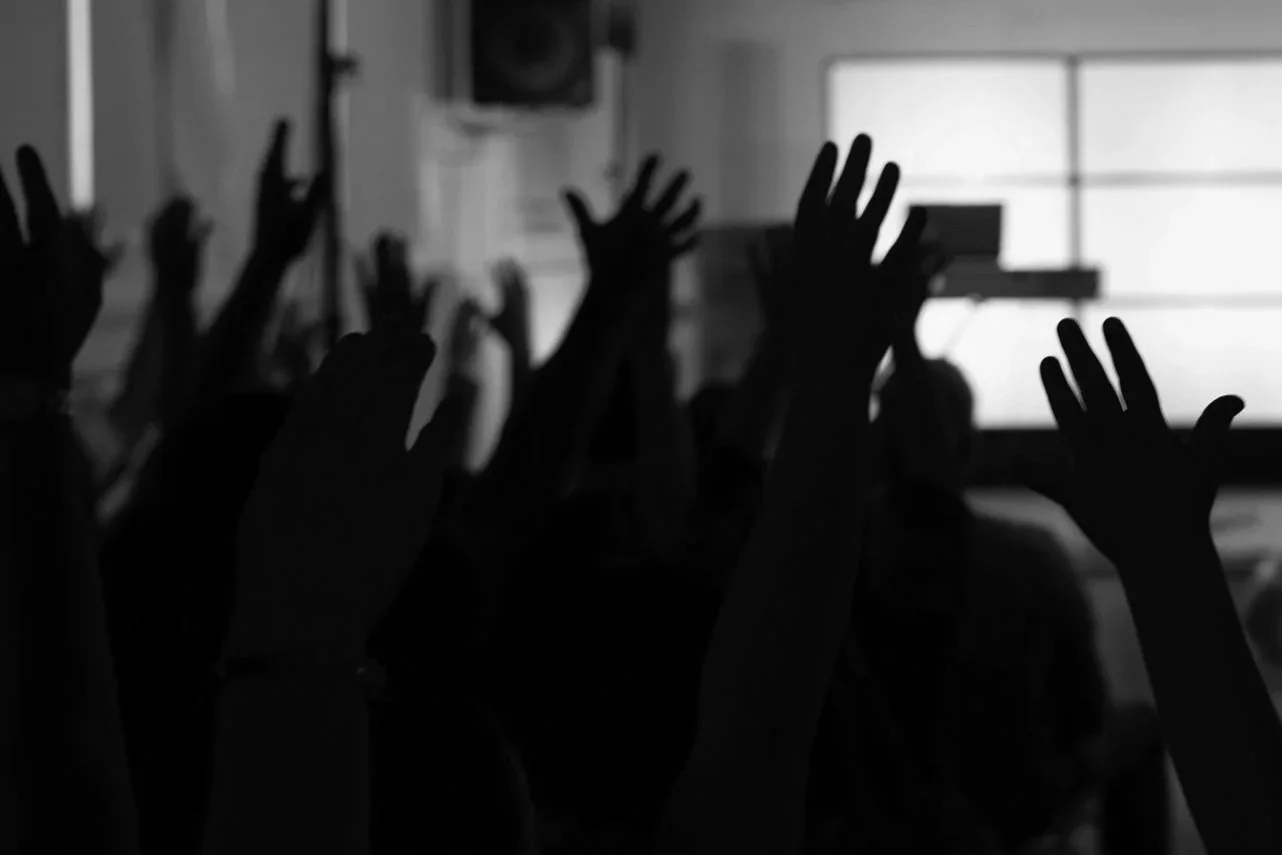 Silhouettes of raised hands in a dark room against bright windows, suggesting a group of people participating in an activity or event.