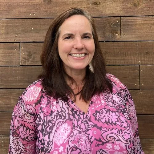 A woman with shoulder-length brown hair smiling, wearing a pink and purple patterned blouse, standing in front of a wooden wall.