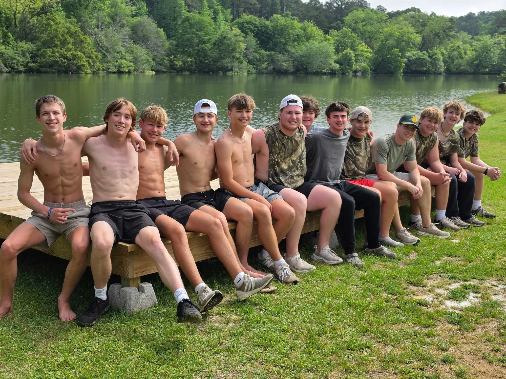 A group of twelve boys sitting on a wooden dock near a lake or river, surrounded by lush green trees, smiling and posing for the photo. Some are shirtless, others wear t-shirts, shorts, and baseball caps.