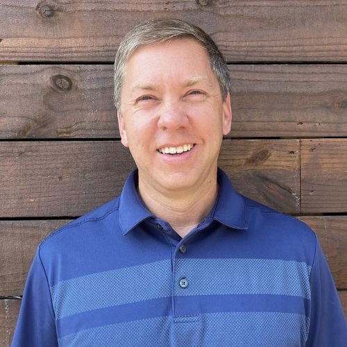 A smiling man with short gray hair wearing a blue polo shirt standing in front of a wooden wall.