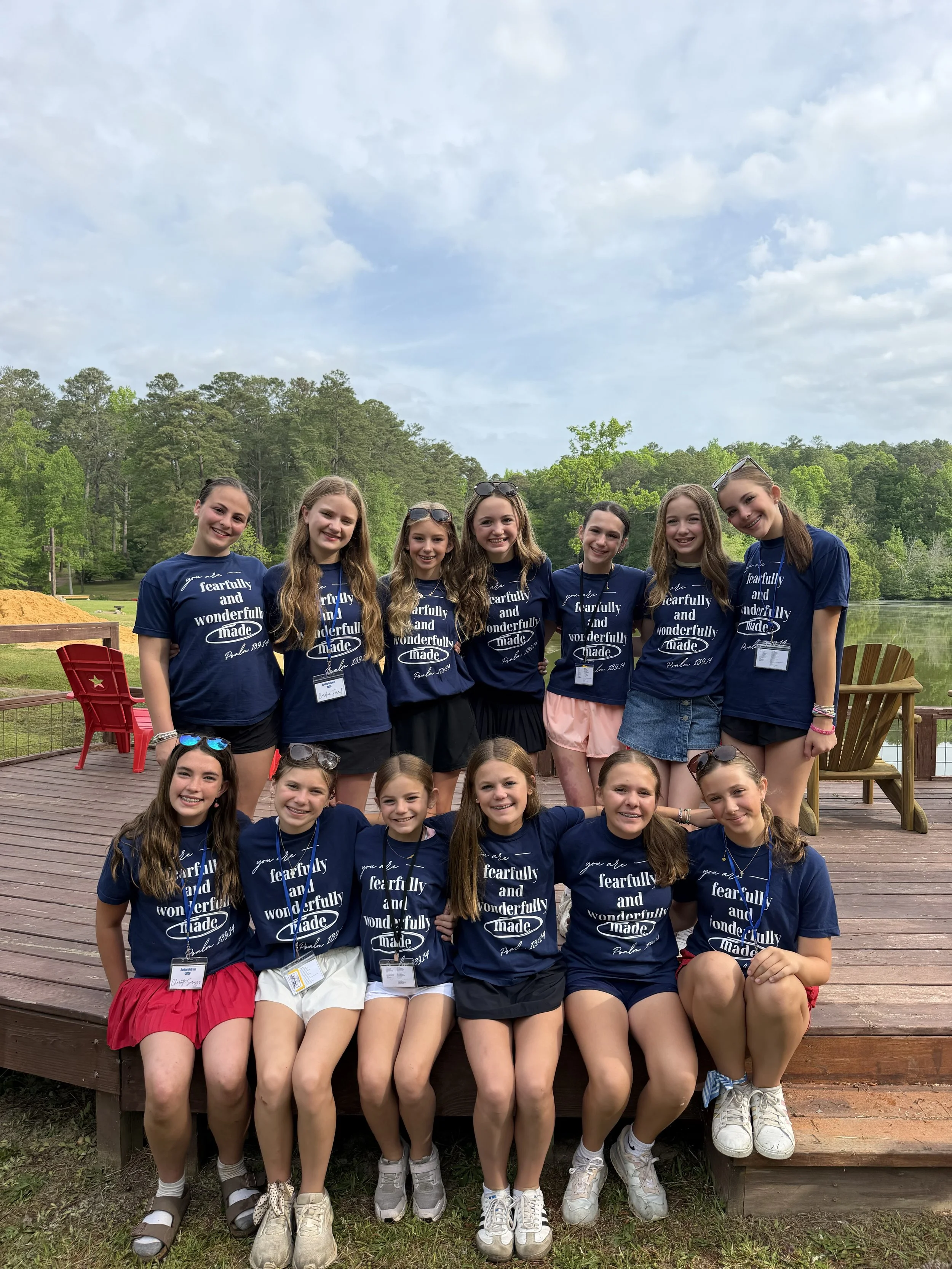 Group of young girls wearing matching navy blue t-shirts with a phrase, standing on a wooden deck outdoors with trees and cloudy sky in the background.