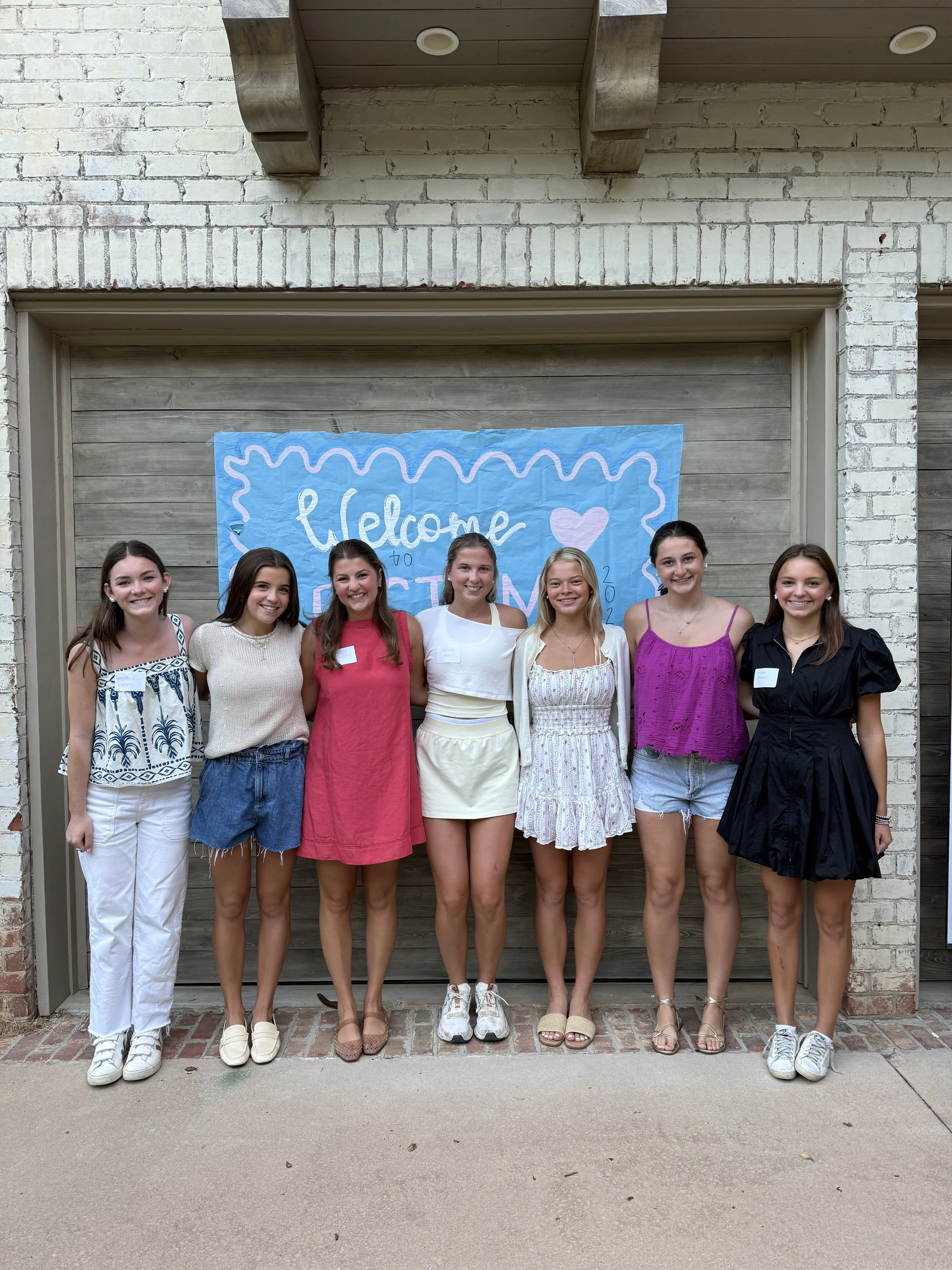 Six young women standing in front of a brick wall with a blue welcome banner, smiling at the camera.