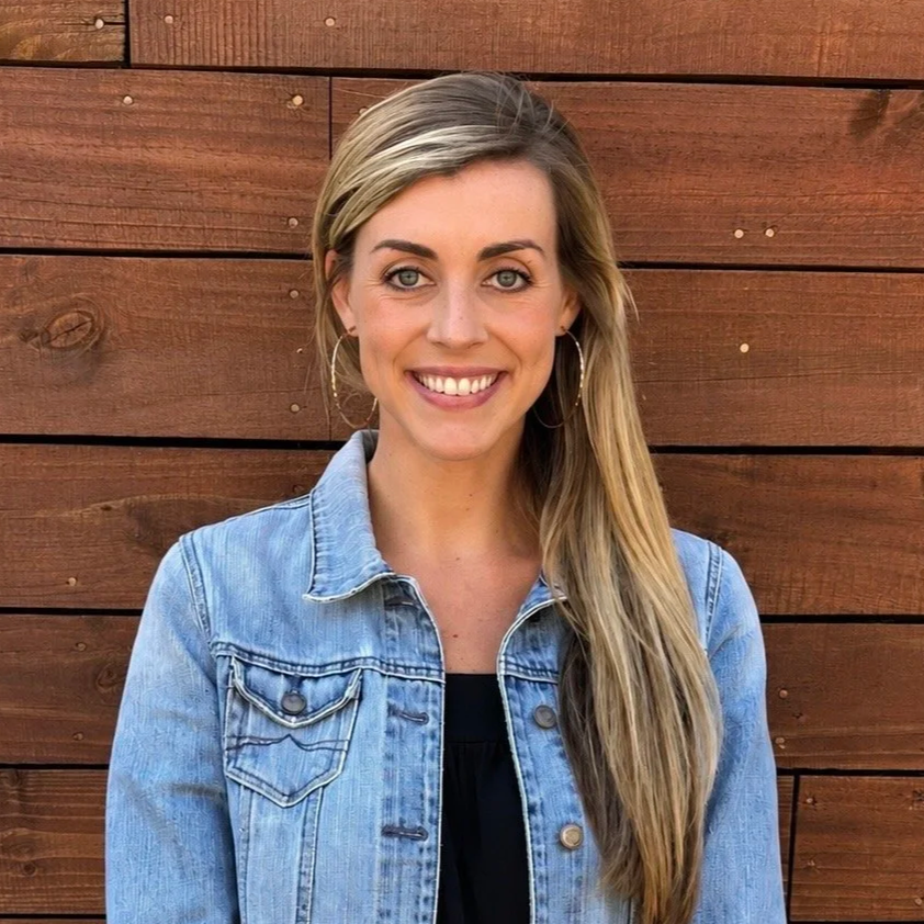 A woman with long blonde hair, wearing hoop earrings, a black top, and a light blue denim jacket, smiling in front of a wooden wall.