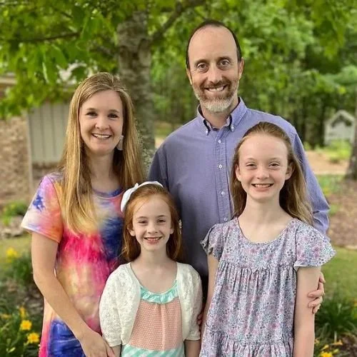 A family of five smiling outdoors in front of green trees. The group consists of two young girls, a young girl, a middle-aged man, and a young woman. They are dressed casually and appear happy.