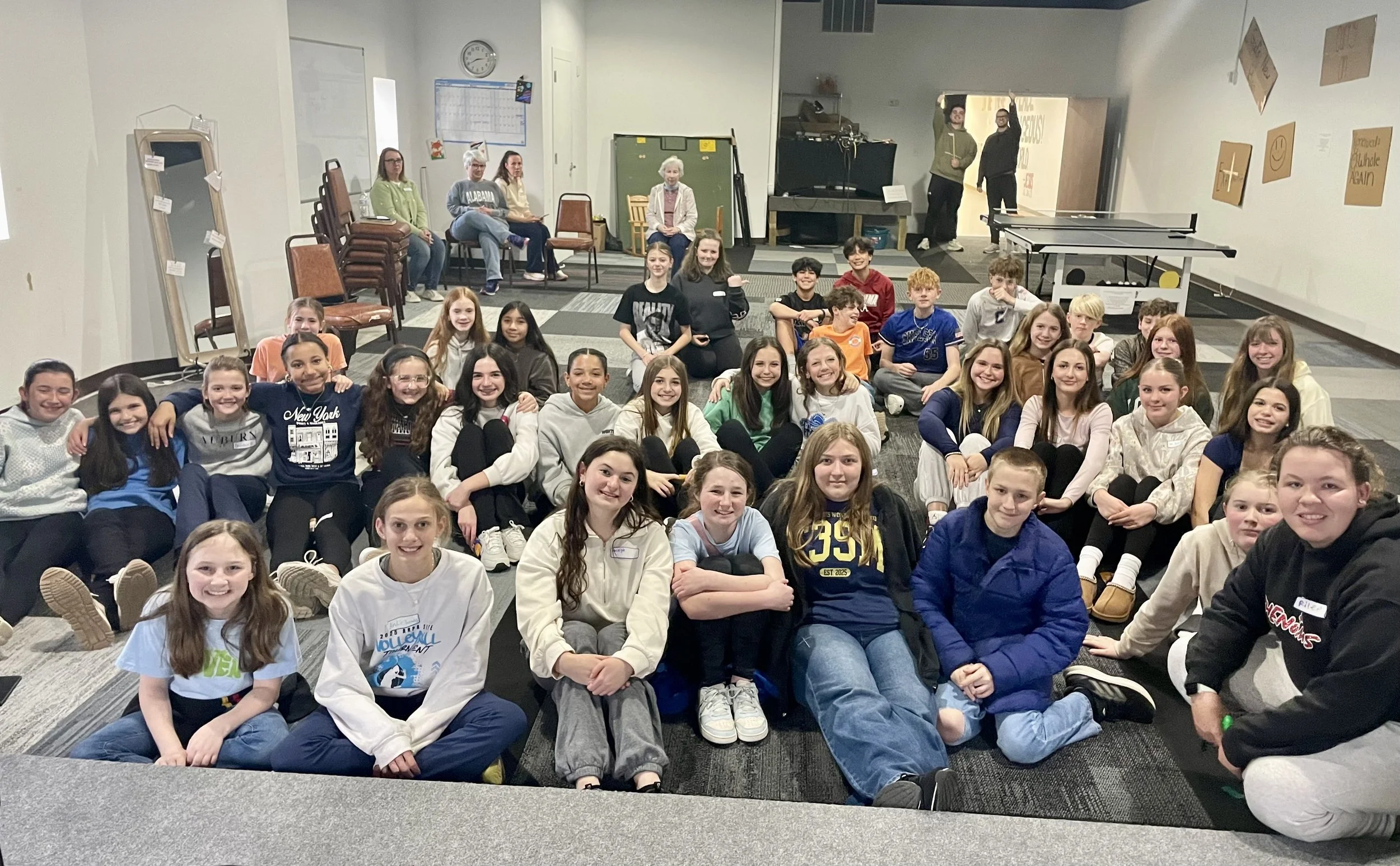 Group of smiling children sitting on the floor in a classroom or event space, with some adults sitting and standing in the background near a door and chalkboard, and a ping pong table on the right.