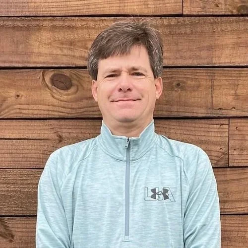 A man standing outdoors in front of a wooden fence, wearing a light blue Under Armour jacket, with a slight smile.