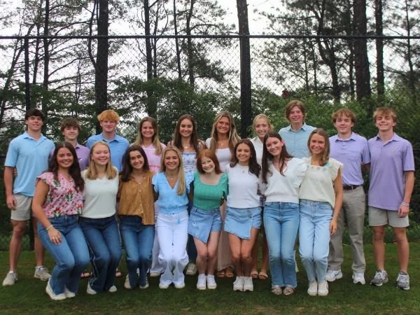 Group of teenagers standing outdoors in front of a chain-link fence and trees, smiling for a photo.