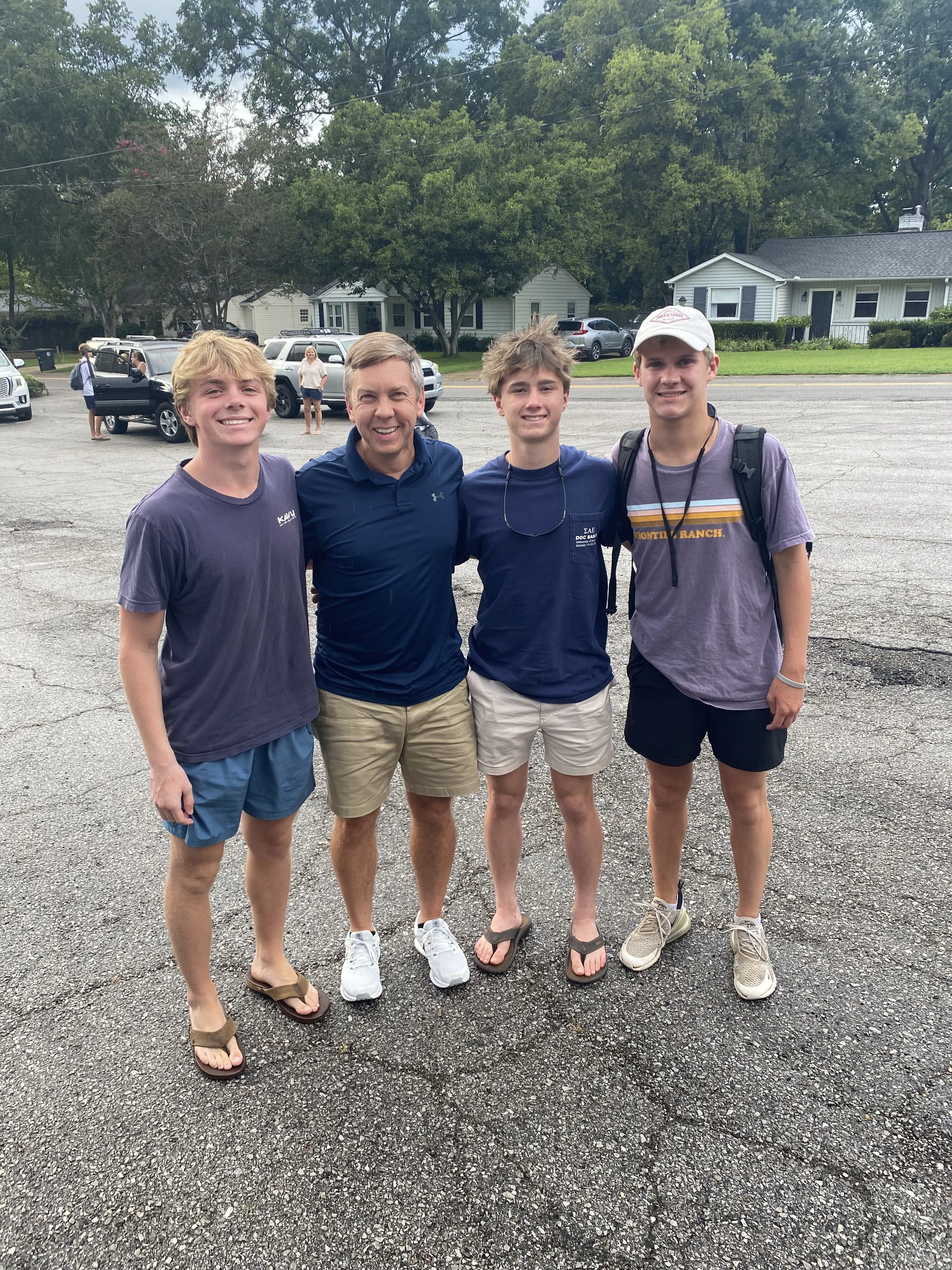 Four young men and one older man standing together outdoors on a cloudy day, smiling. They are dressed casually in T-shirts, shorts, and sandals or sneakers. Behind them, there are houses, trees, and parked cars.