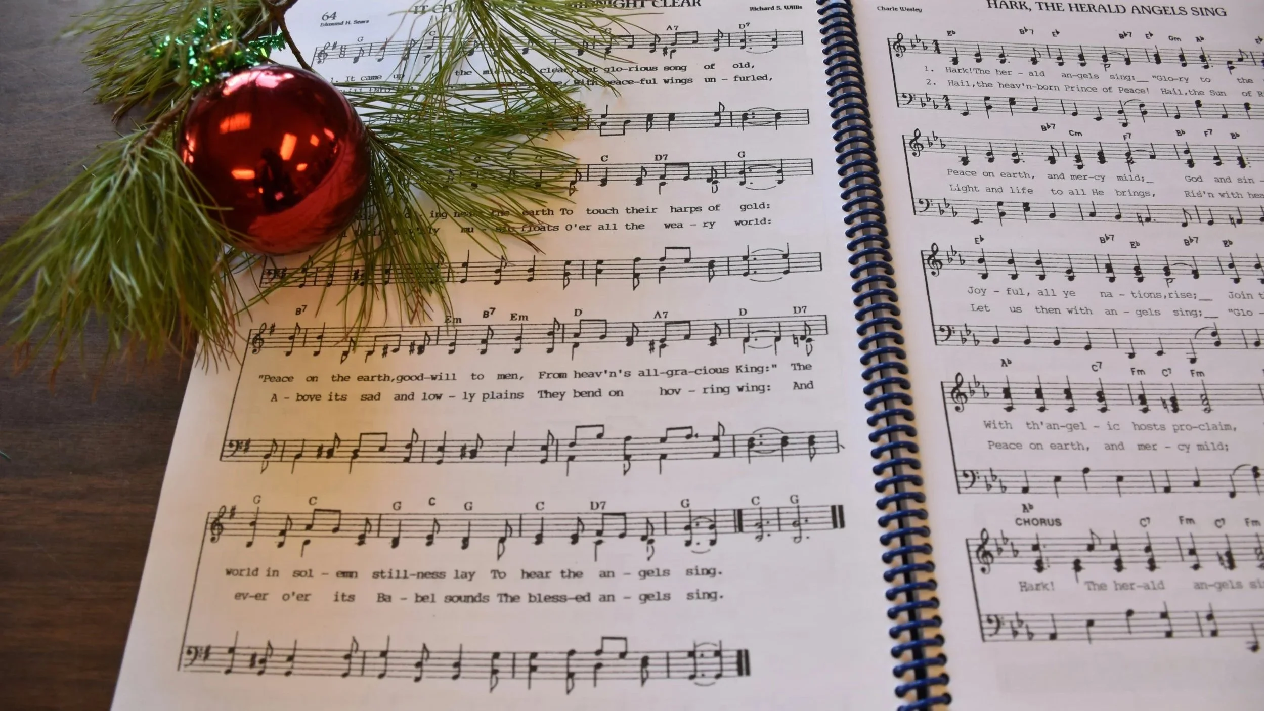 A photograph of sheet music adorned with a red Christmas tree ornament and an evergreen branch