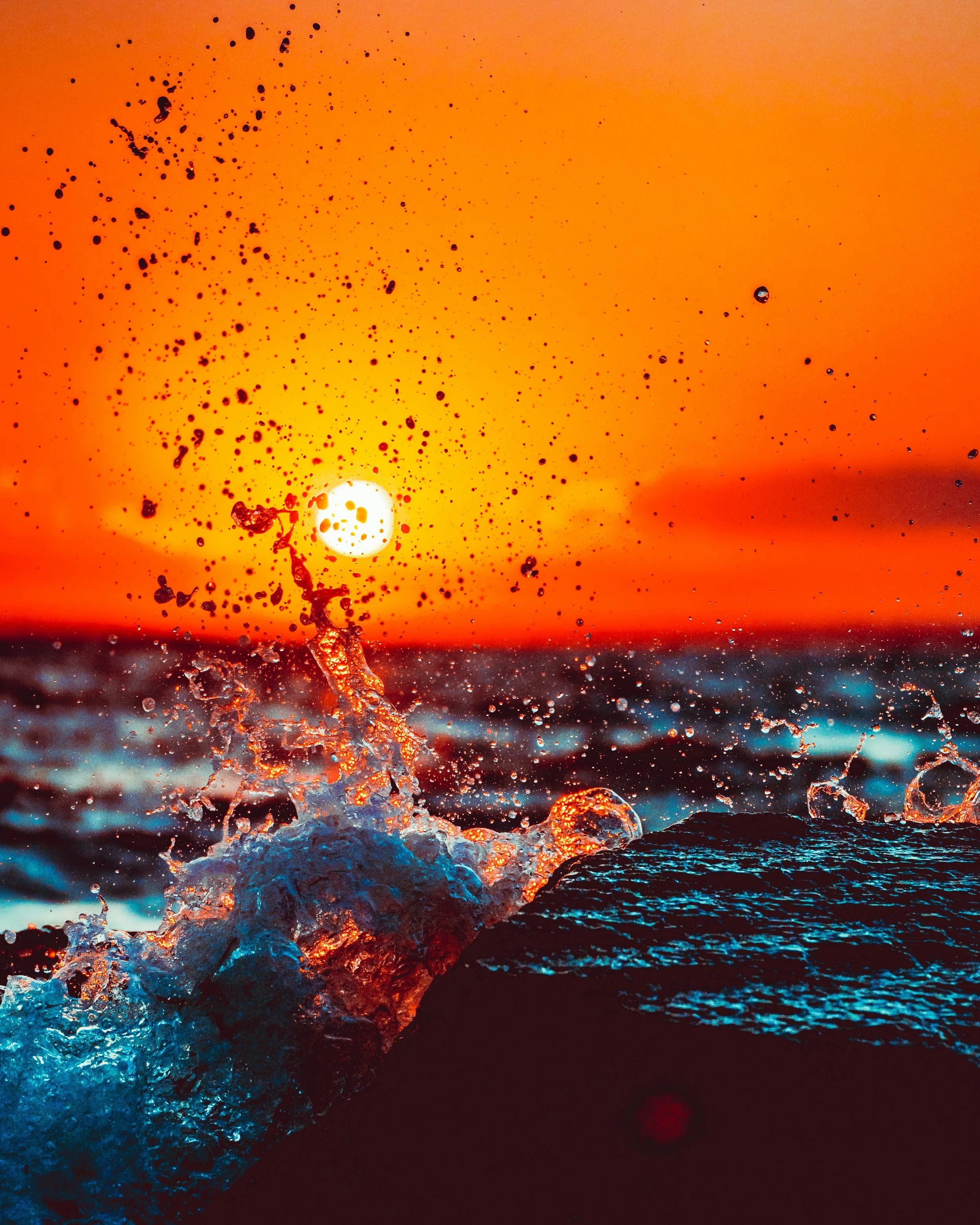 A photo of ocean water splashing up in front of a setting or rising sun